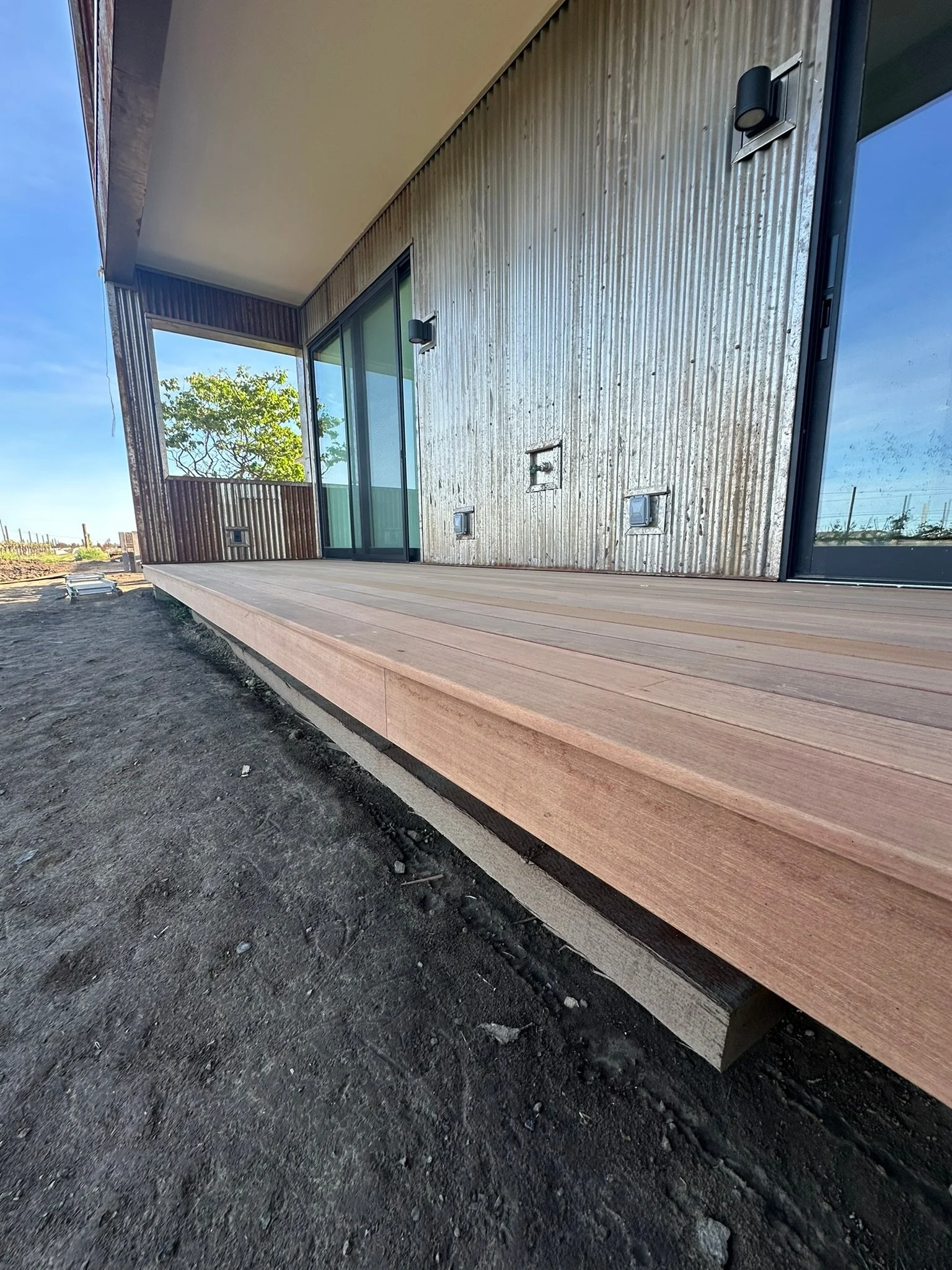 Exterior view of a modern house with a wooden deck, sliding glass doors, and black exterior lights, with a tree and clear sky in the background.