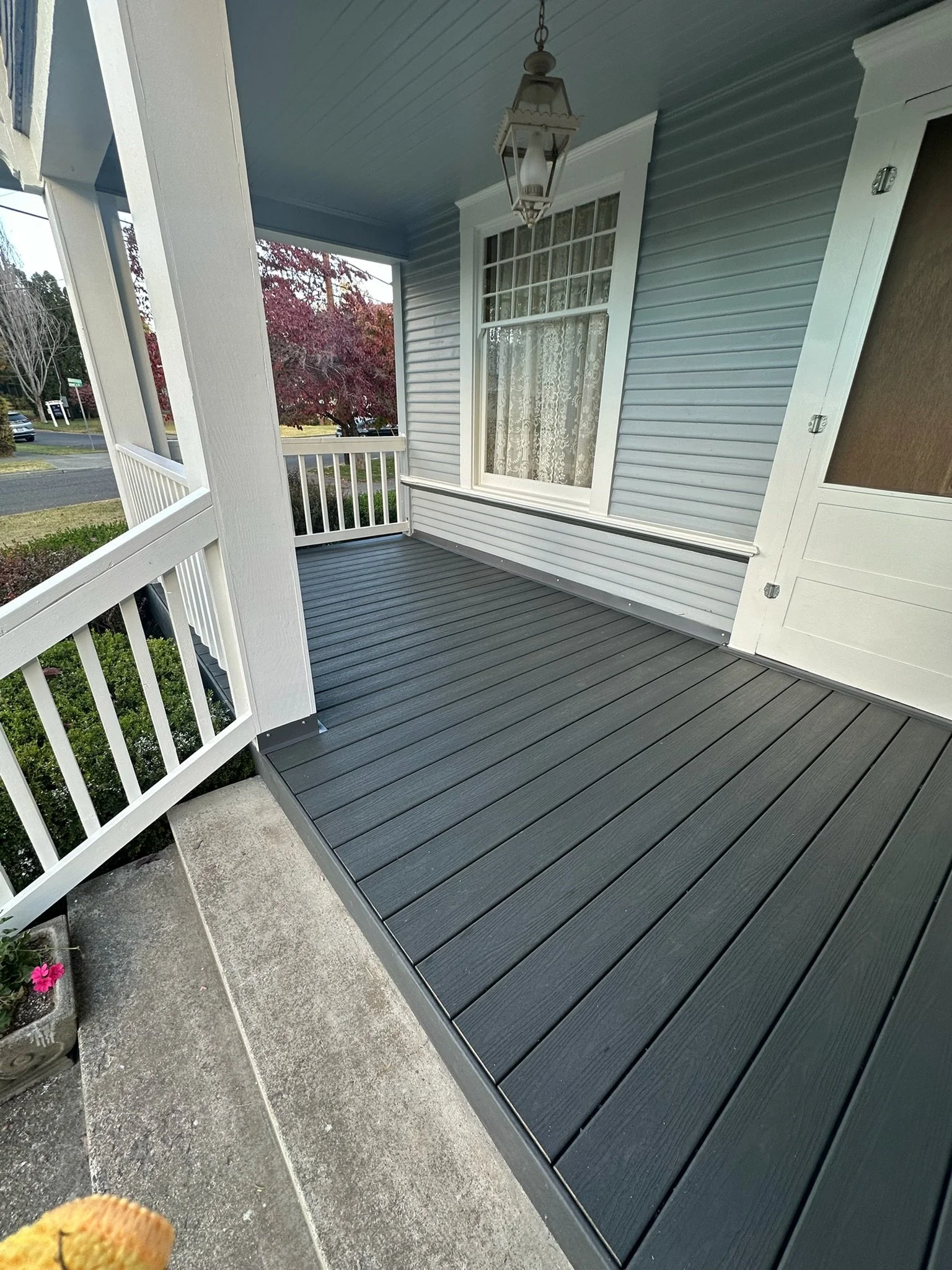 View of a porch with gray flooring, white railing, and a house with light blue siding and white trim. A window with lace curtains and a hanging lantern light fixture are visible. Some pink flowers are in a pot near the porch.