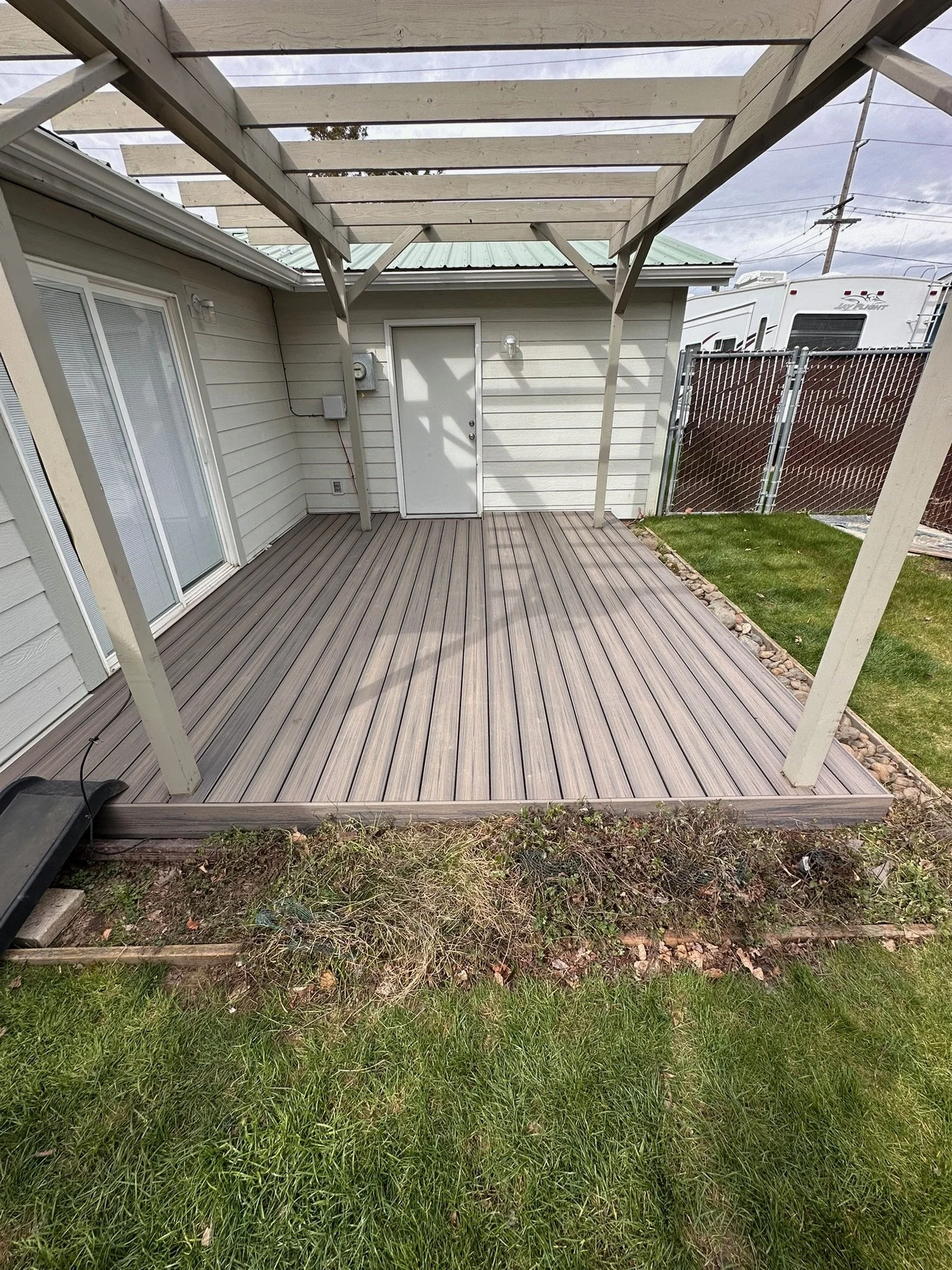 Backyard patio with a partially built wooden pergola, a sliding glass door, an electrical panel, and a small patch of grass with rocks beside the patio.