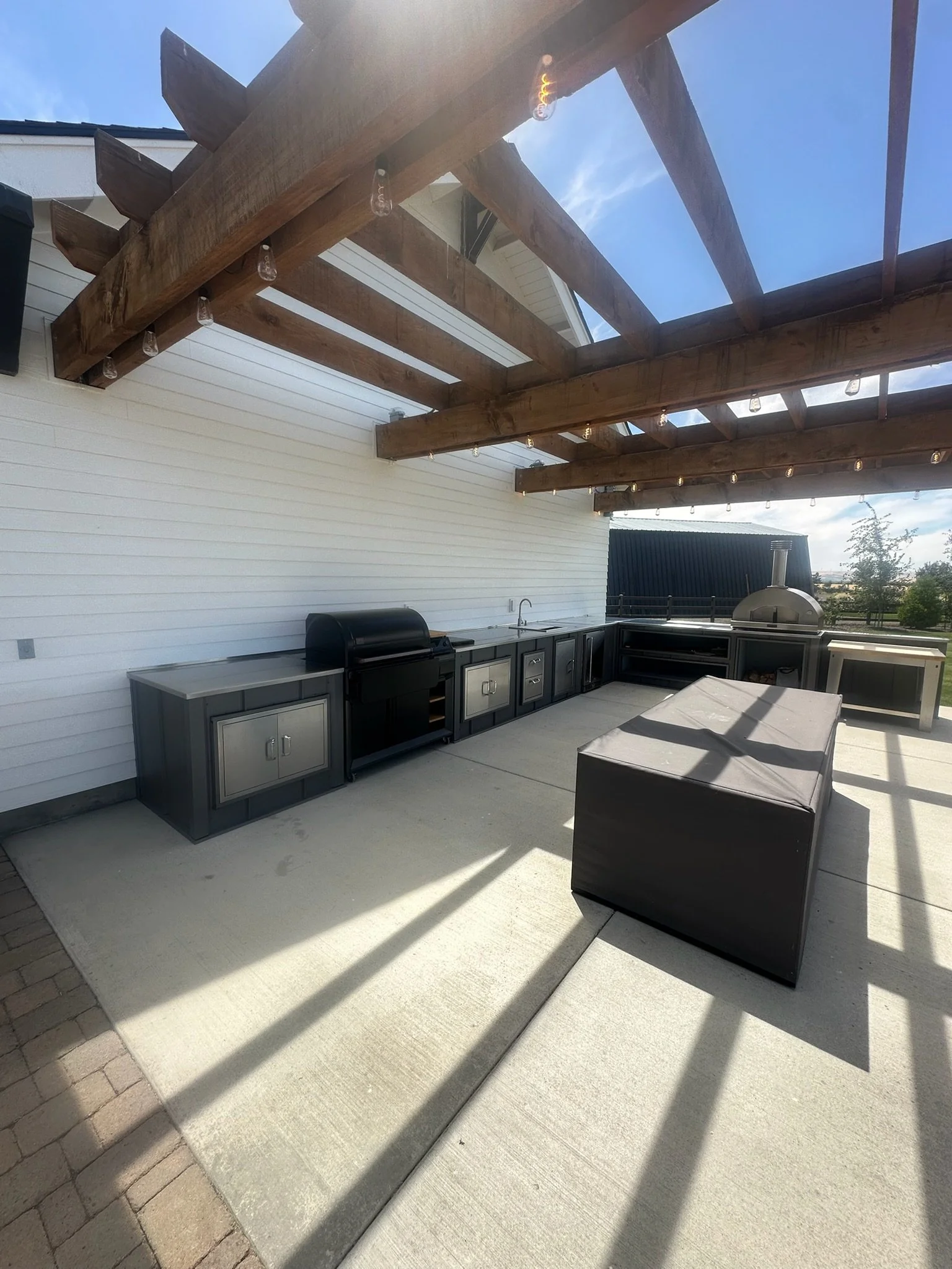 An outdoor patio area with a white wall and black cabinets beneath a wooden pergola, which has string lights hanging from it. There is a black grill and a covered outdoor oven or smoker, with a patio table in the foreground. Shadows are cast on the concrete and brick flooring.