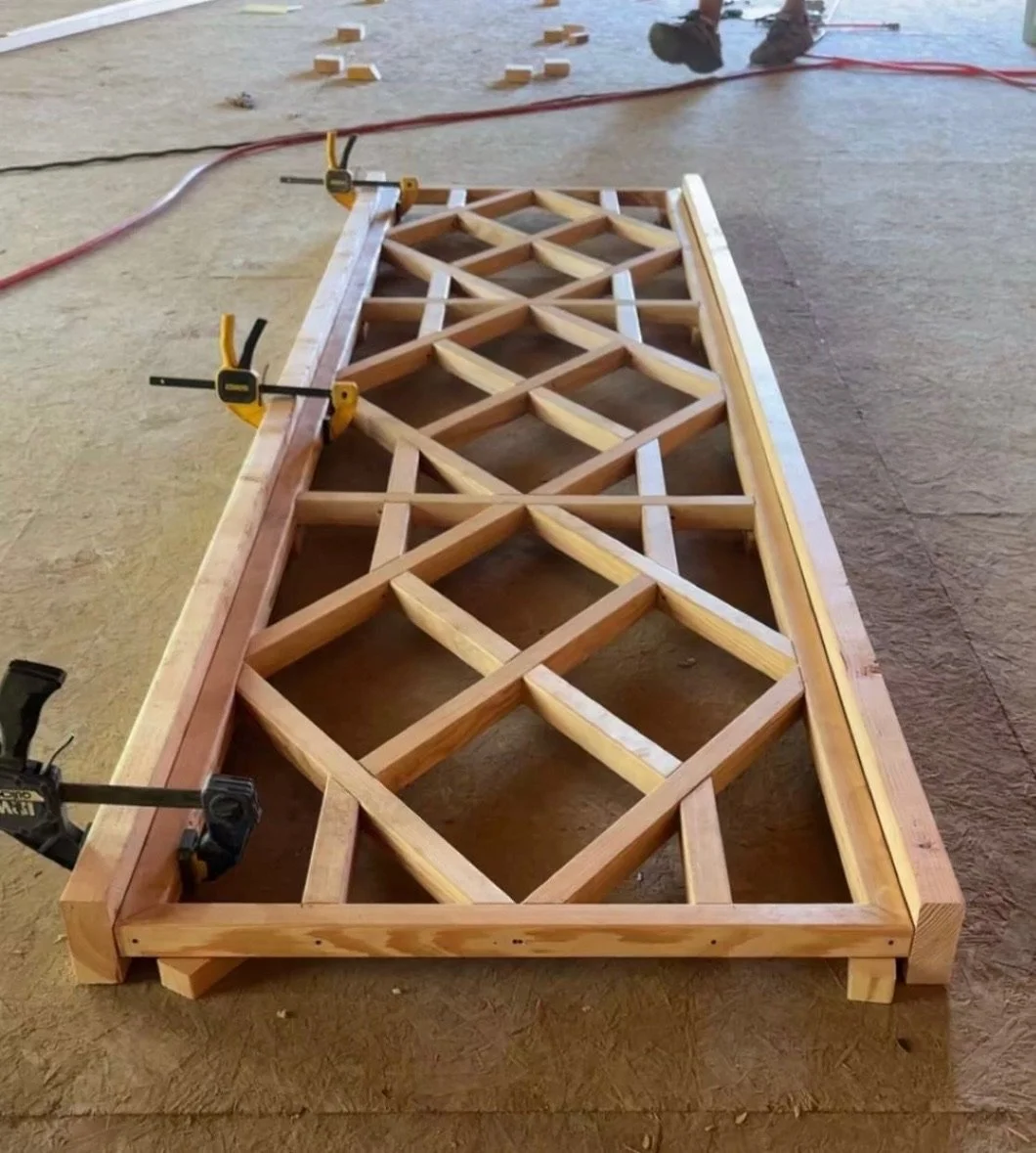A wooden truss bridge frame being assembled on a work surface with clamps securing the pieces together.