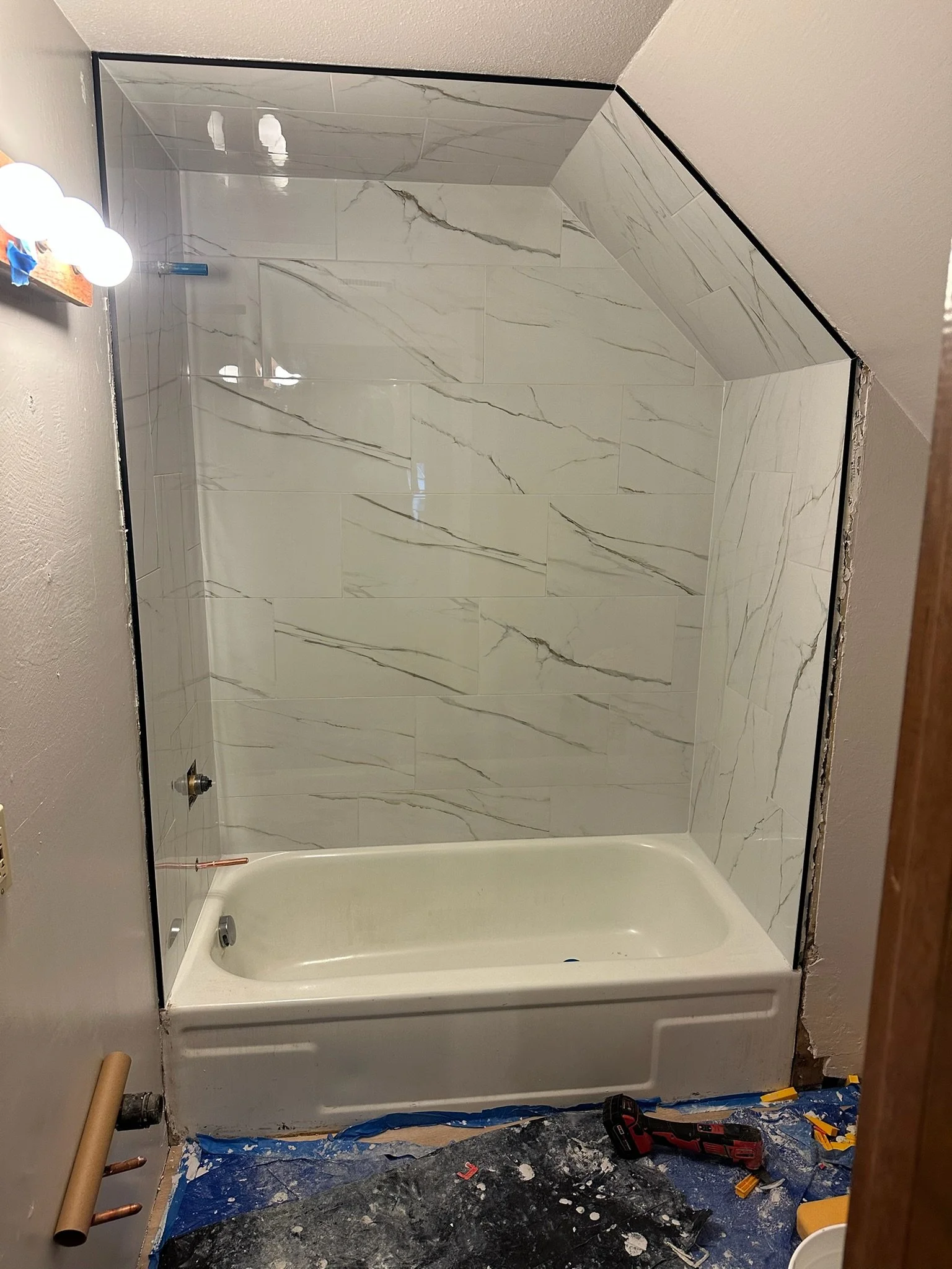 Bathroom renovation in progress with a new marble tile shower installed. Construction tools, a blue drop cloth, and debris on the floor indicate ongoing work.