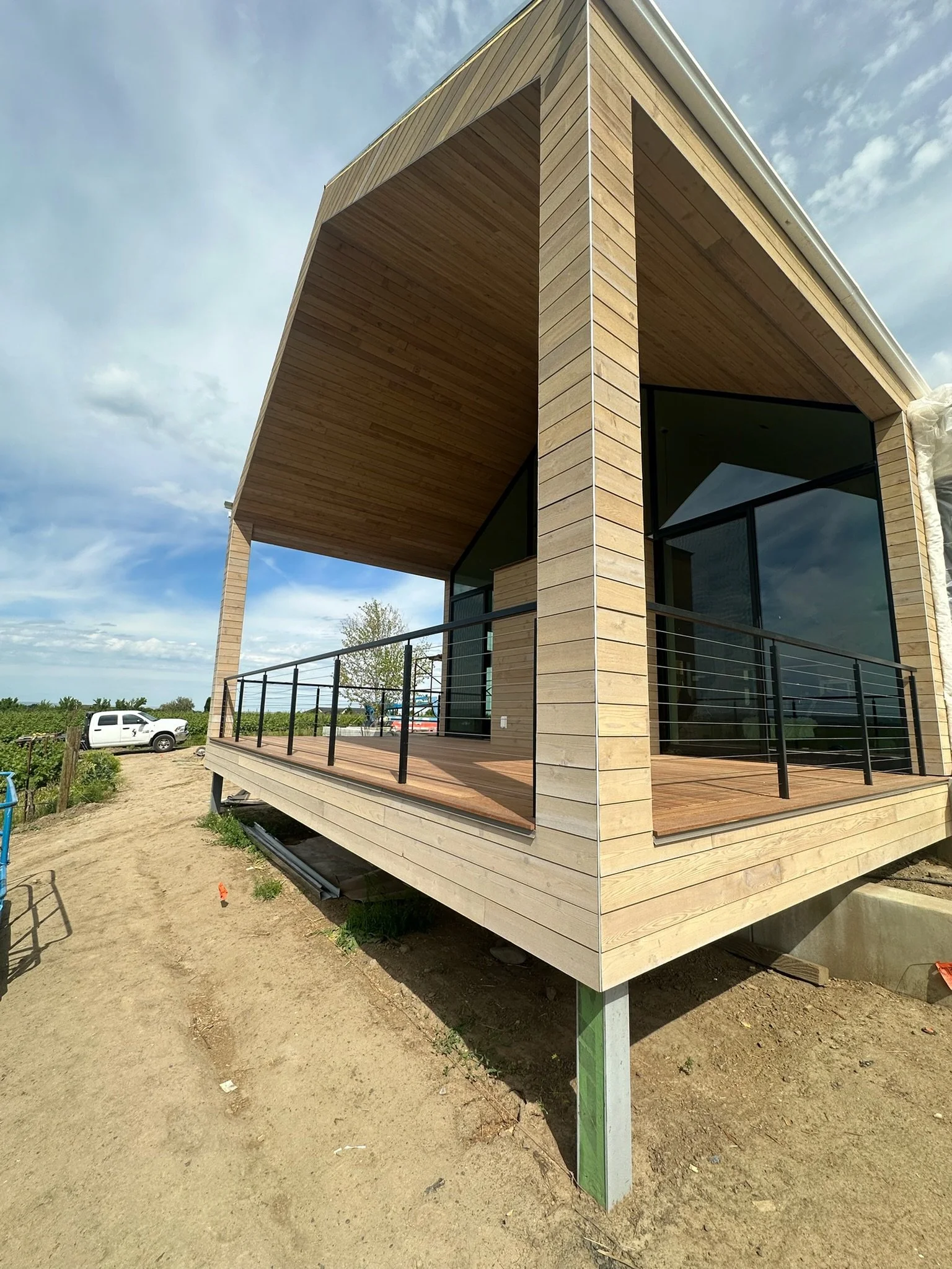 A modern house with a light wood exterior, large glass windows, and a spacious balcony with a black railing. The house is situated outdoors with a partly cloudy sky and a white vehicle parked nearby.