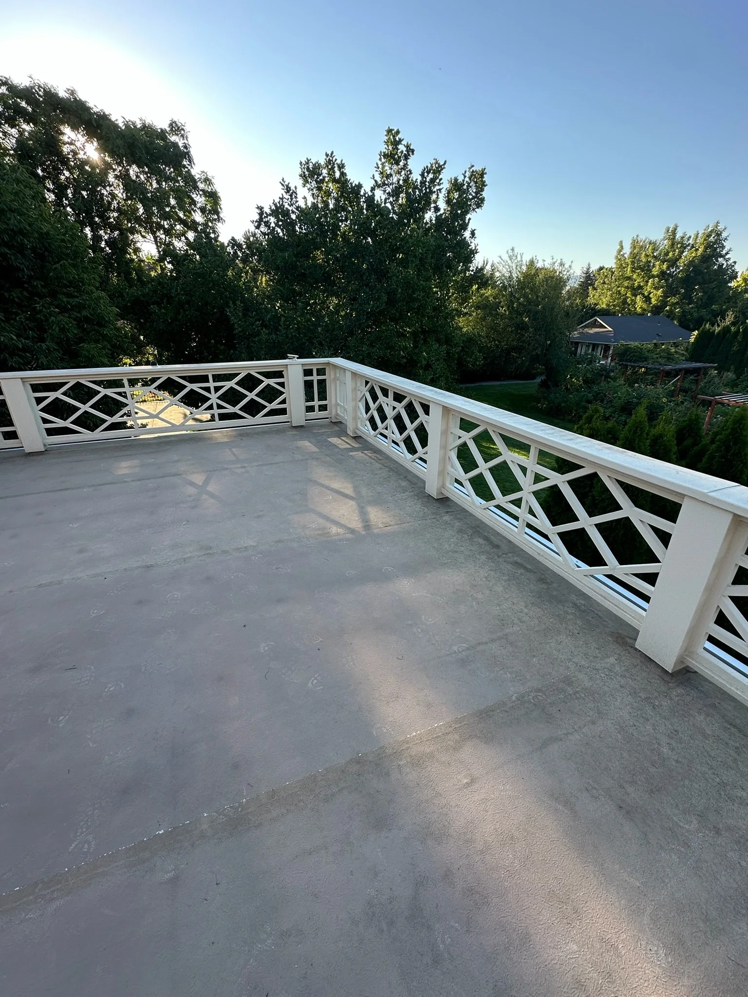 An outdoor balcony with a white decorative railing, a concrete floor, and trees in the background under a clear blue sky.