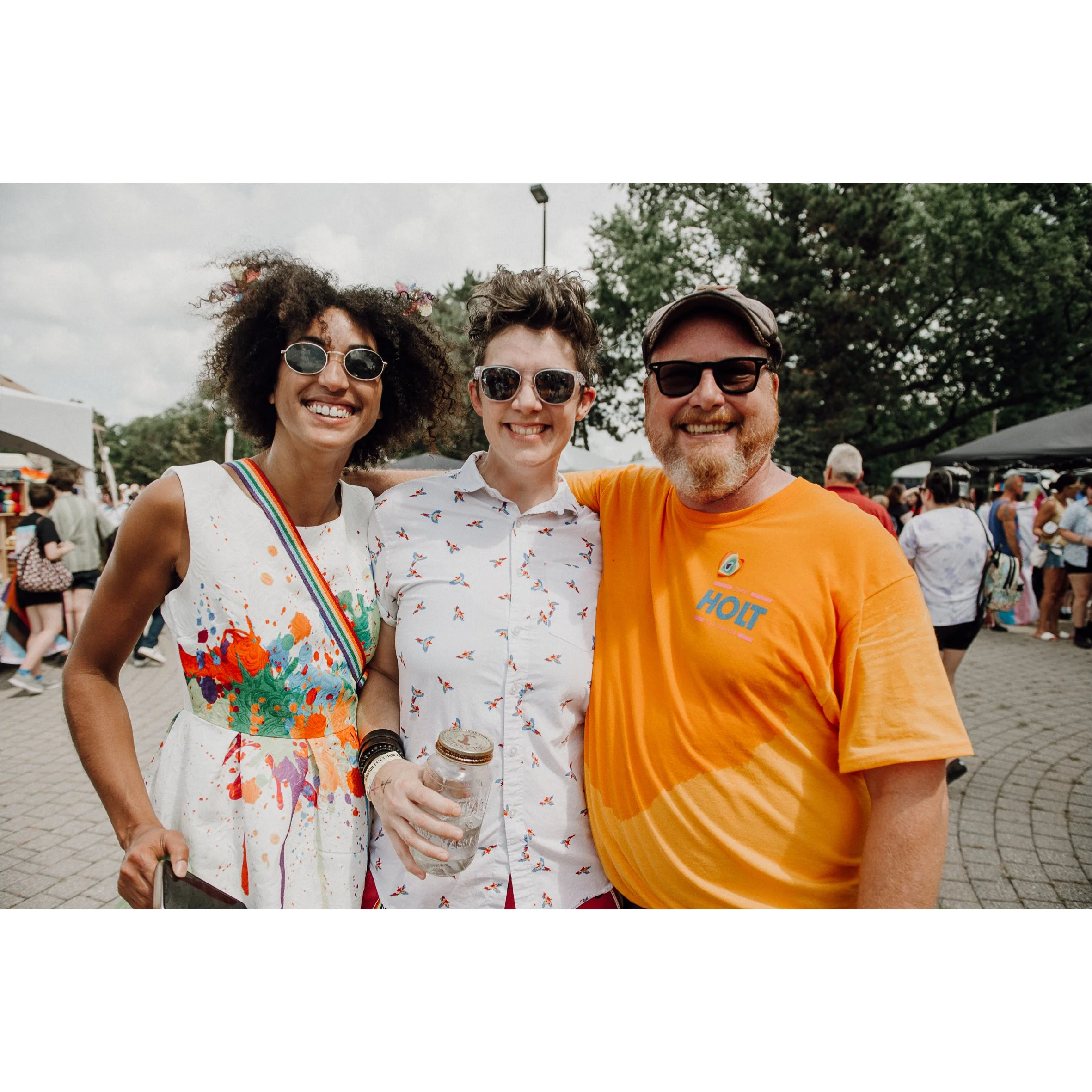 Three smiling people are fitting together for a group photo at an outdoor festival or event. The woman on the left has curly hair, sunglasses, and a white dress with colorful paint splatters and rainbow straps. The man in the middle has short hair, glasses, and a shirt with birds on it. The man on the right has a beard, glasses, a cap, and wears an orange shirt with a logo that says 'HOLT.'
