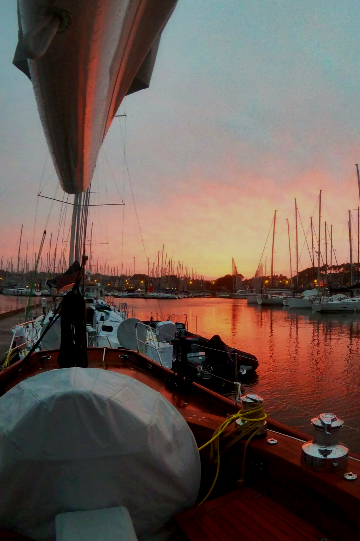 View of a marina at sunset with boats docked, calm water reflecting the orange and pink sky, and a sailboat mast in the foreground.