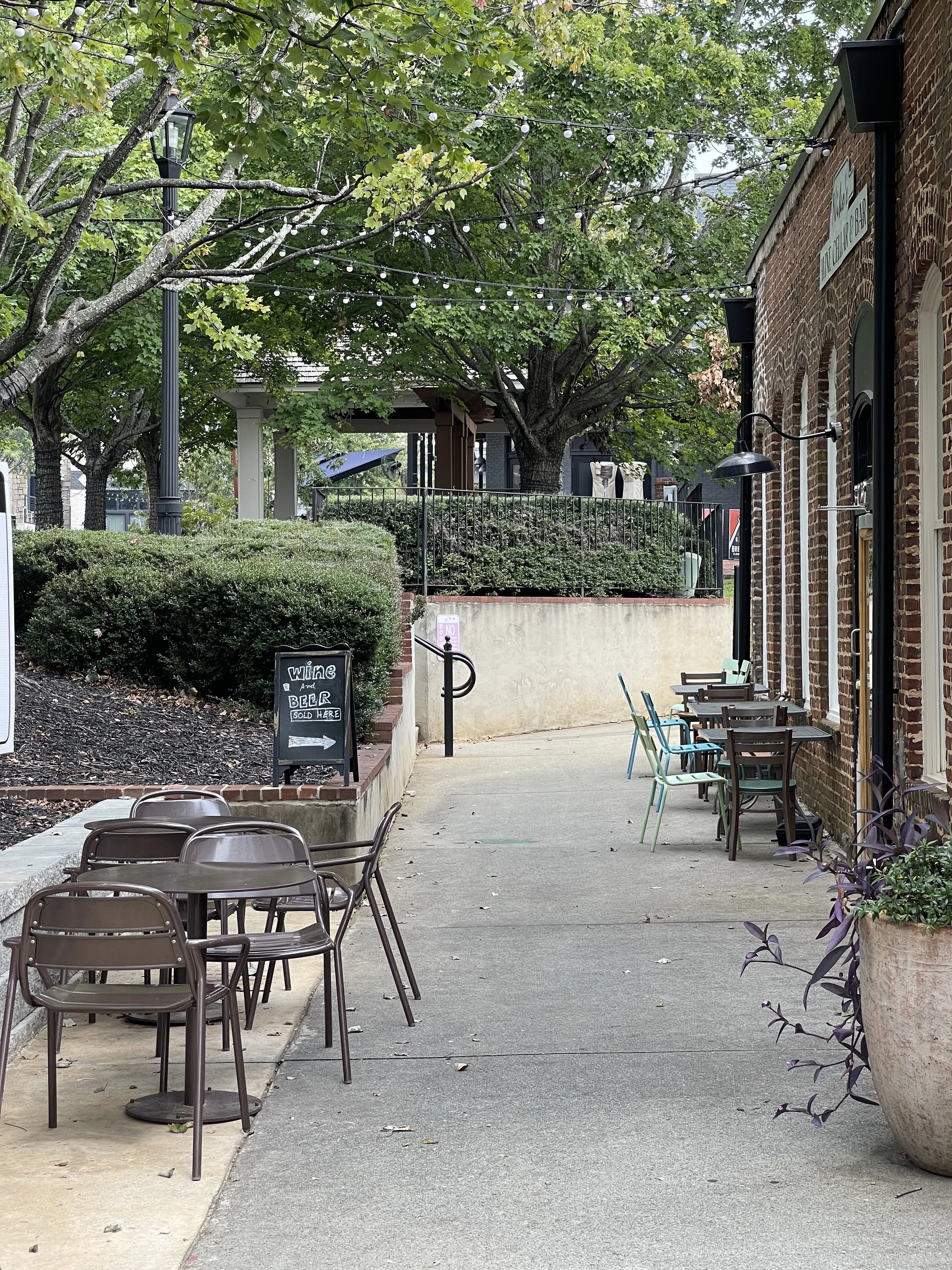 Empty outdoor seating area of a restaurant with tables and chairs, a sign indicating wine and beer sold there, lush green trees with string lights, brick building on the right, sidewalk, and planters with plants.