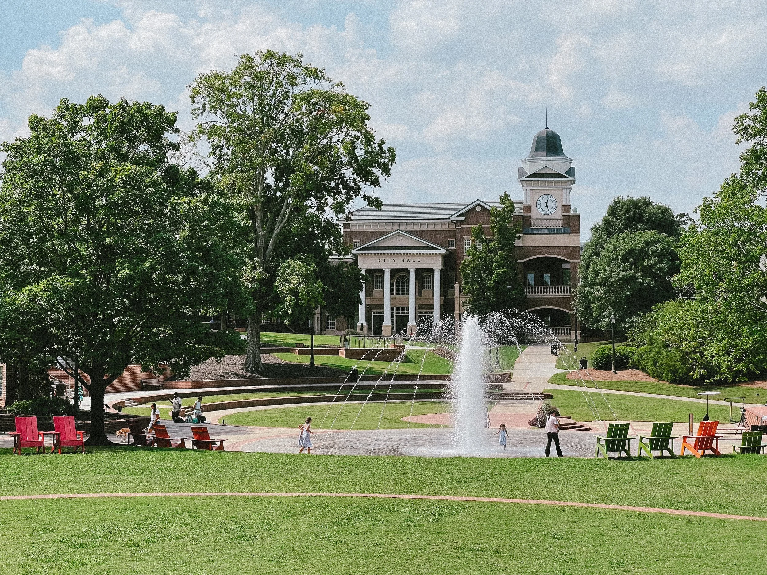 A city park with green grass, trees, and a fountain in the foreground. People are walking and sitting on benches. In the background, there is a large brick city hall building with a clock tower, and a blue sky with some clouds.