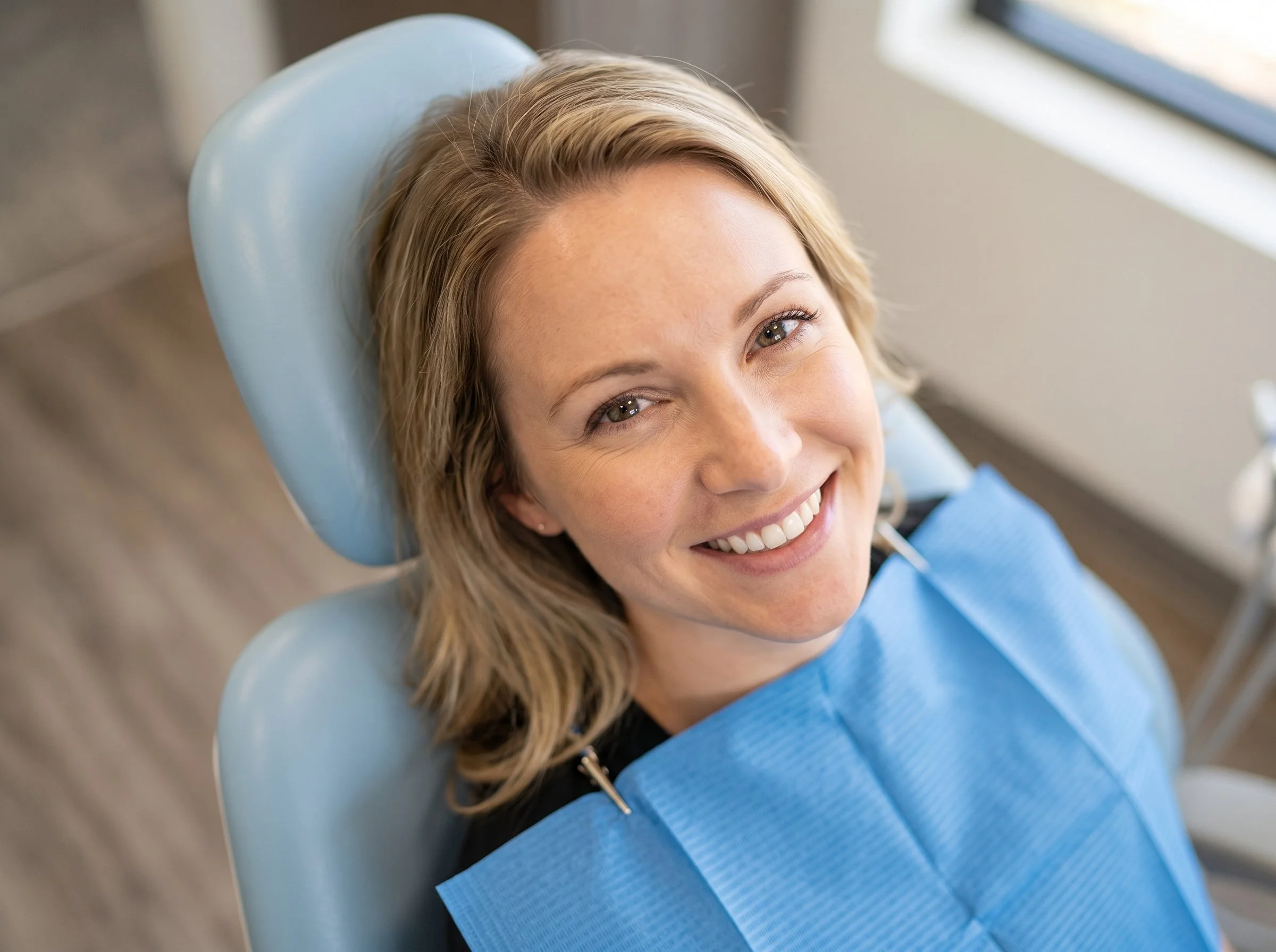 A smiling woman with blonde hair lying back in a dental chair wearing a blue dental gown.