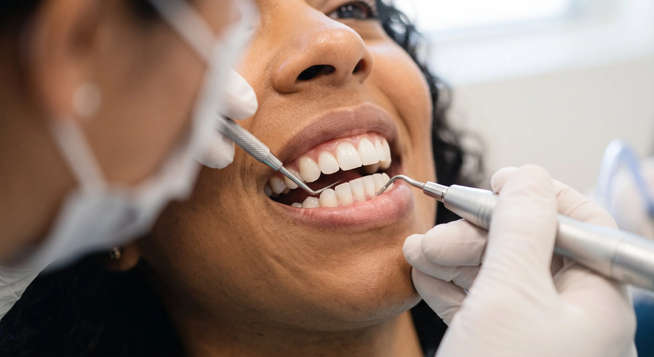 A woman receiving dental cleaning from a dental professional, who is using dental tools while the woman smiles.