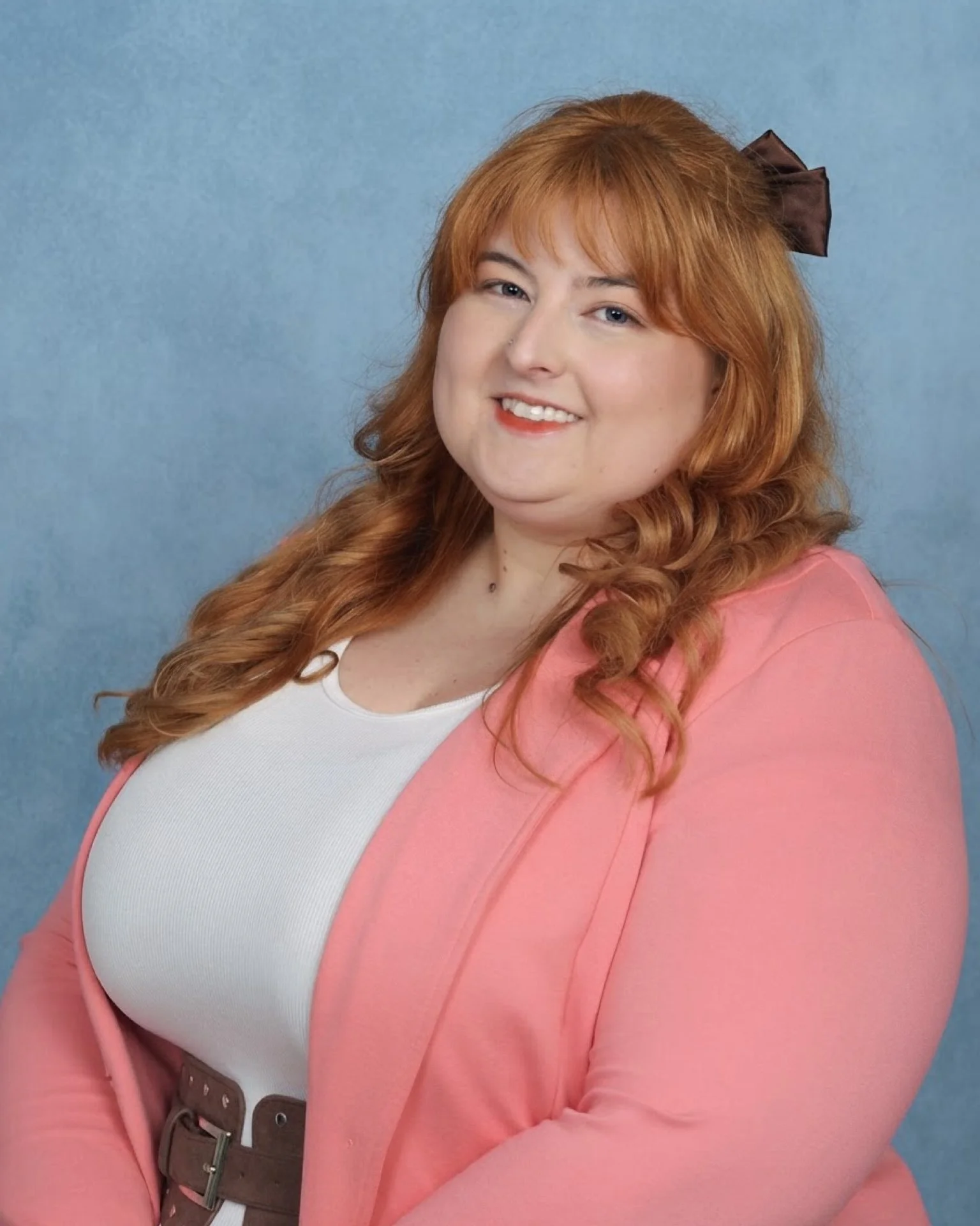 A woman with long wavy red hair wearing a pink blazer over a white top, standing against a light blue background, smiling at the camera.