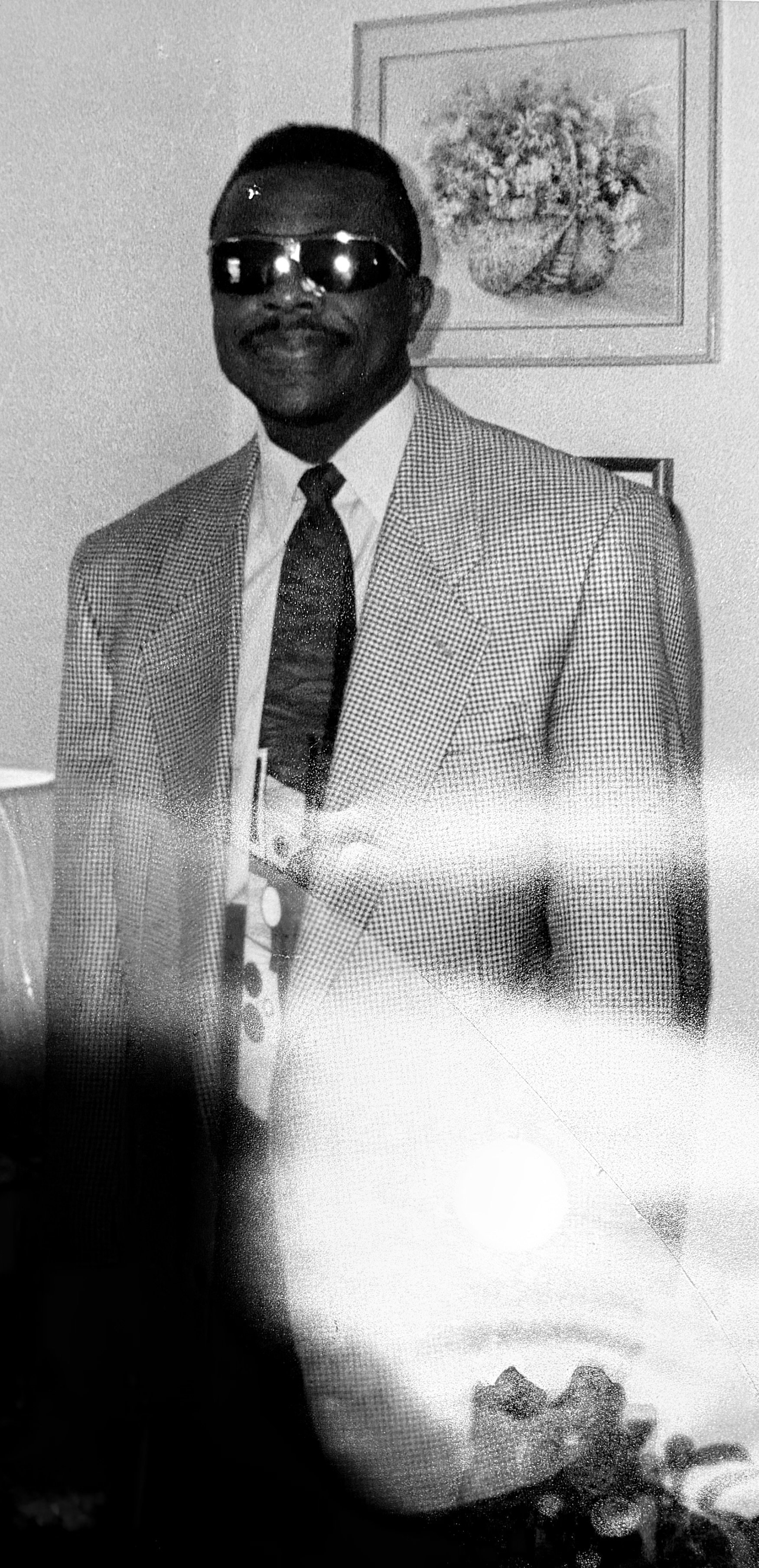 A man wearing sunglasses, a checkered blazer, a white shirt, and a dark tie standing indoors next to a framed floral painting.