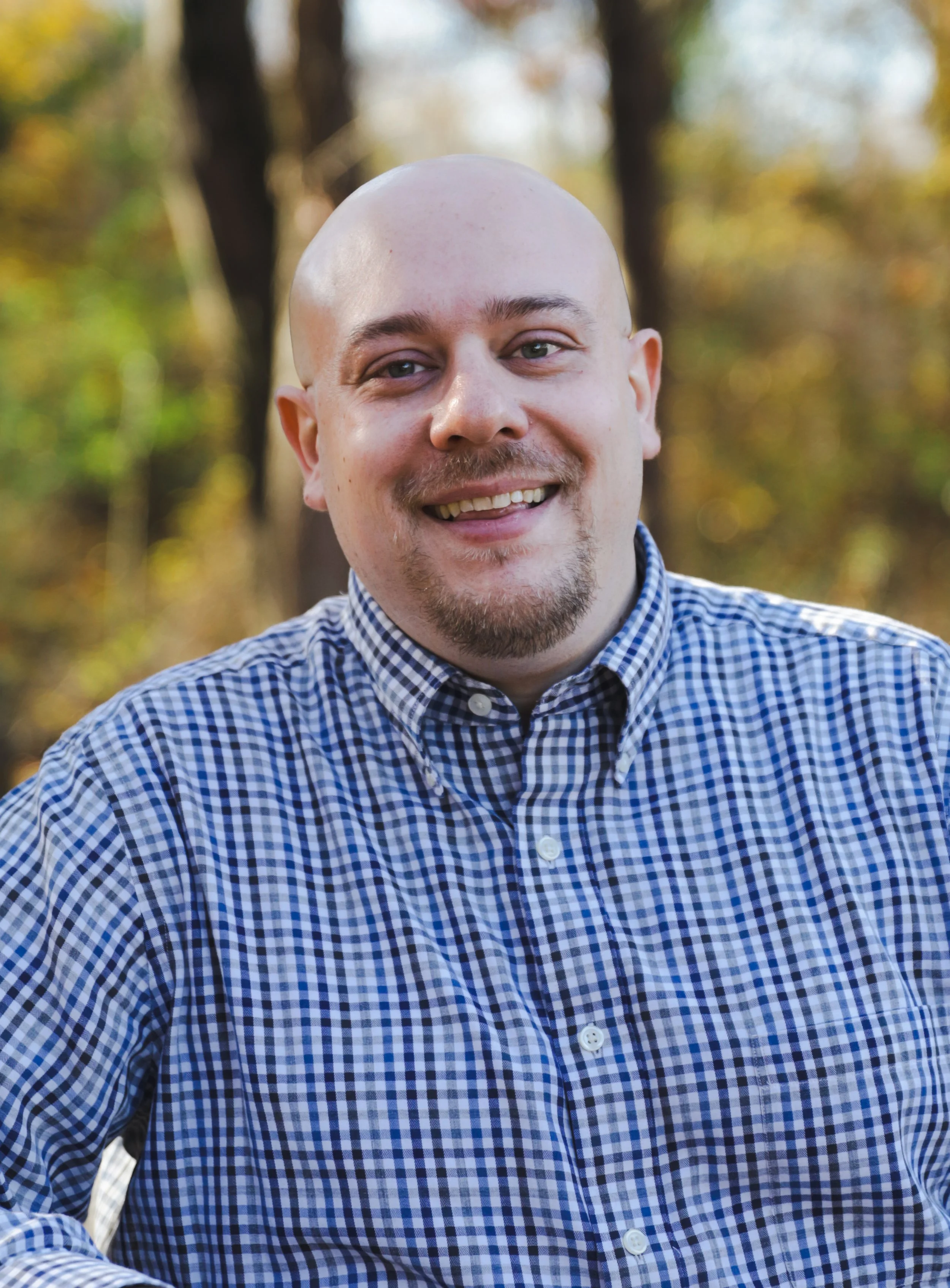 A man with a shaved head and goatee smiling outdoors, wearing a blue and white checkered shirt, with blurred trees and autumn foliage in the background.