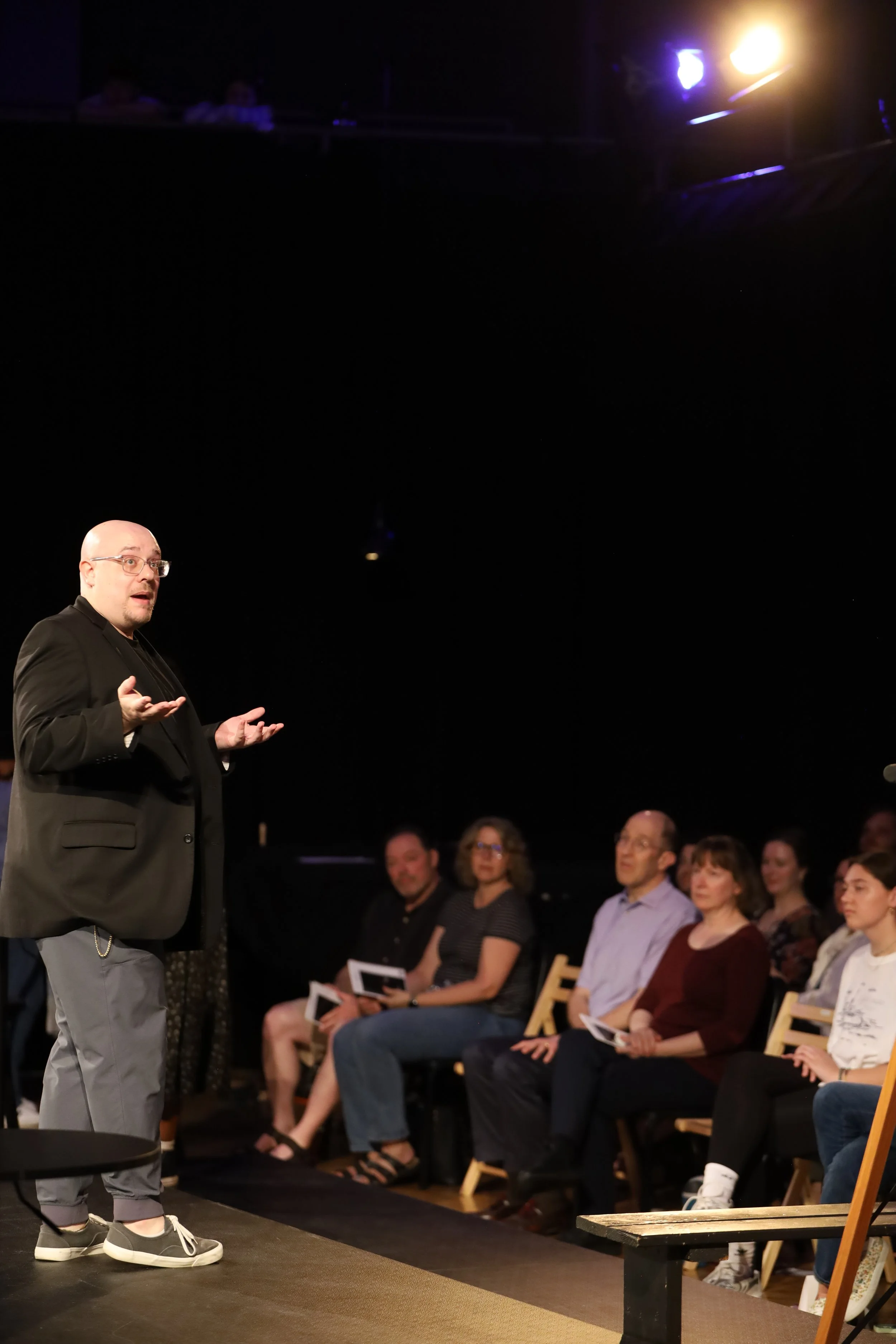A man in a black blazer and grey pants standing on a stage, speaking to an audience of seated adults, with a dark background and stage lighting.