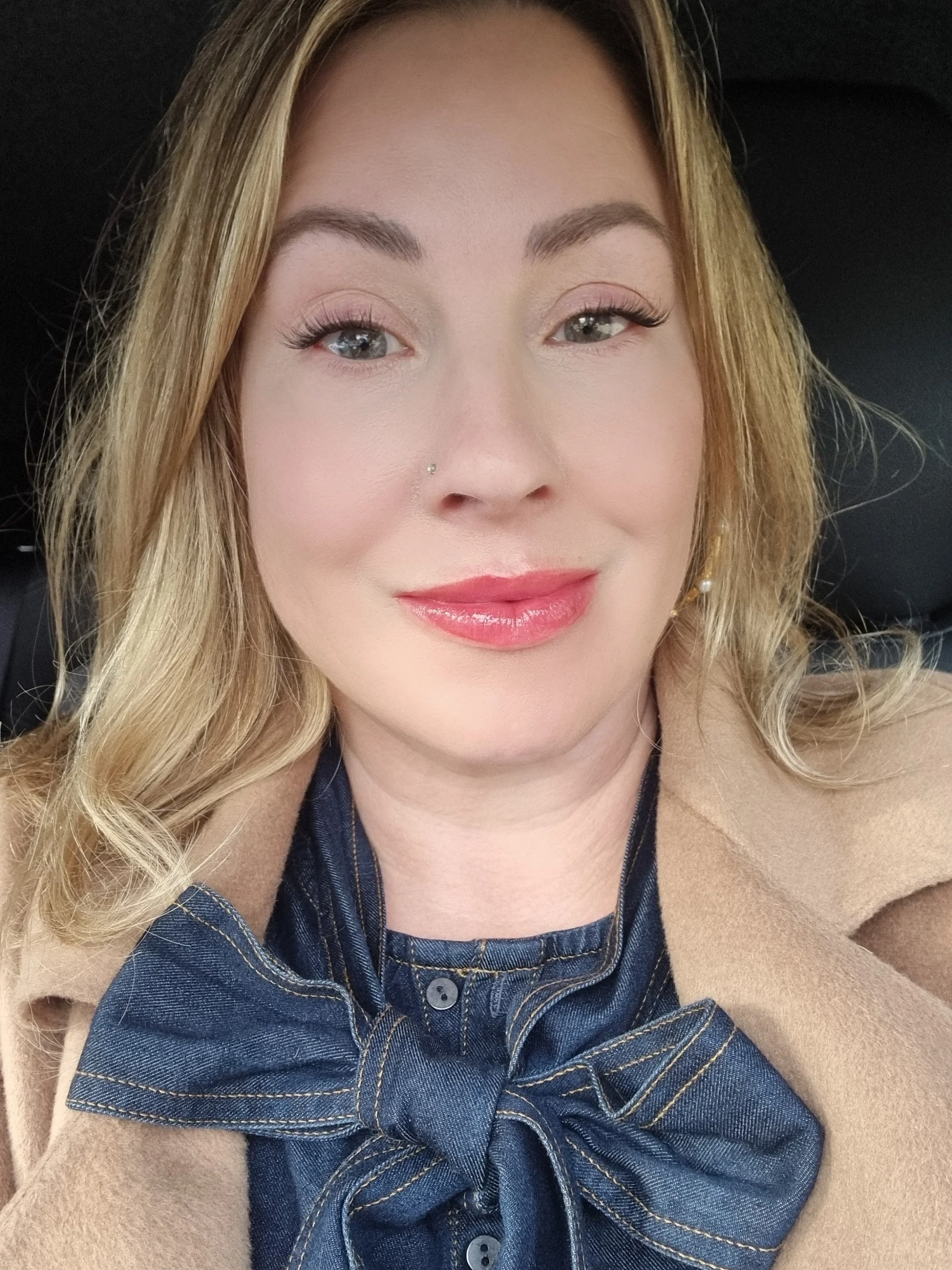 Close-up photo of a woman personal stylist with blonde hair, wearing a beige coat, denim pussy-bow shirt and gold jewellery, smiling openly.