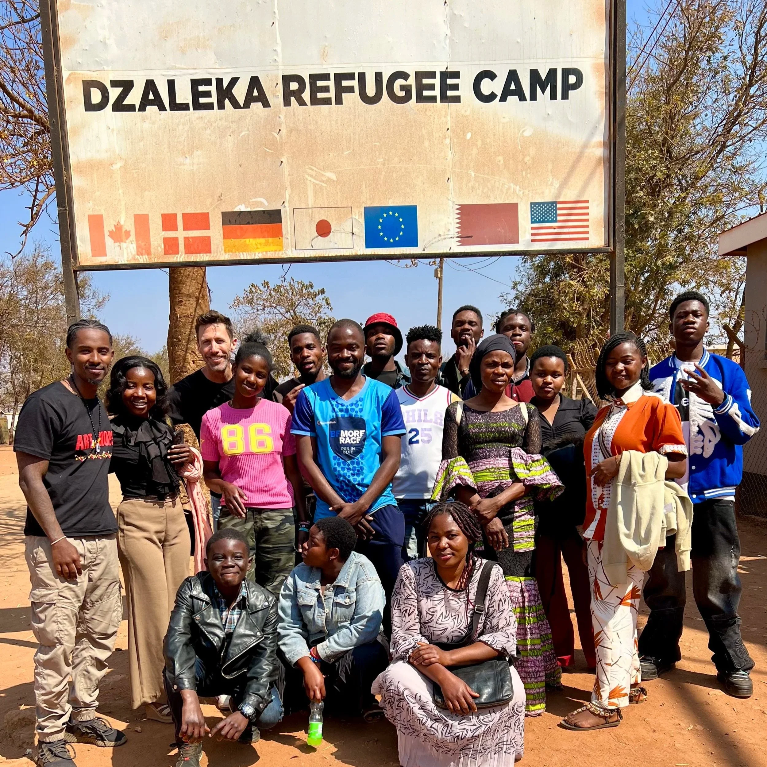 The staff of Dzaleka Refugee Camp's yoga studio and programing standing outdoors in front of a sign that reads "Dzaleka Refugee Camp".