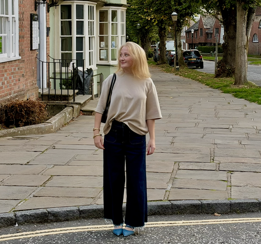 Emily, the Founder of The Conscious Press communications studio, standing outside looking to the side. She is wearing navy jeans, a beige top and blue kitten heels.