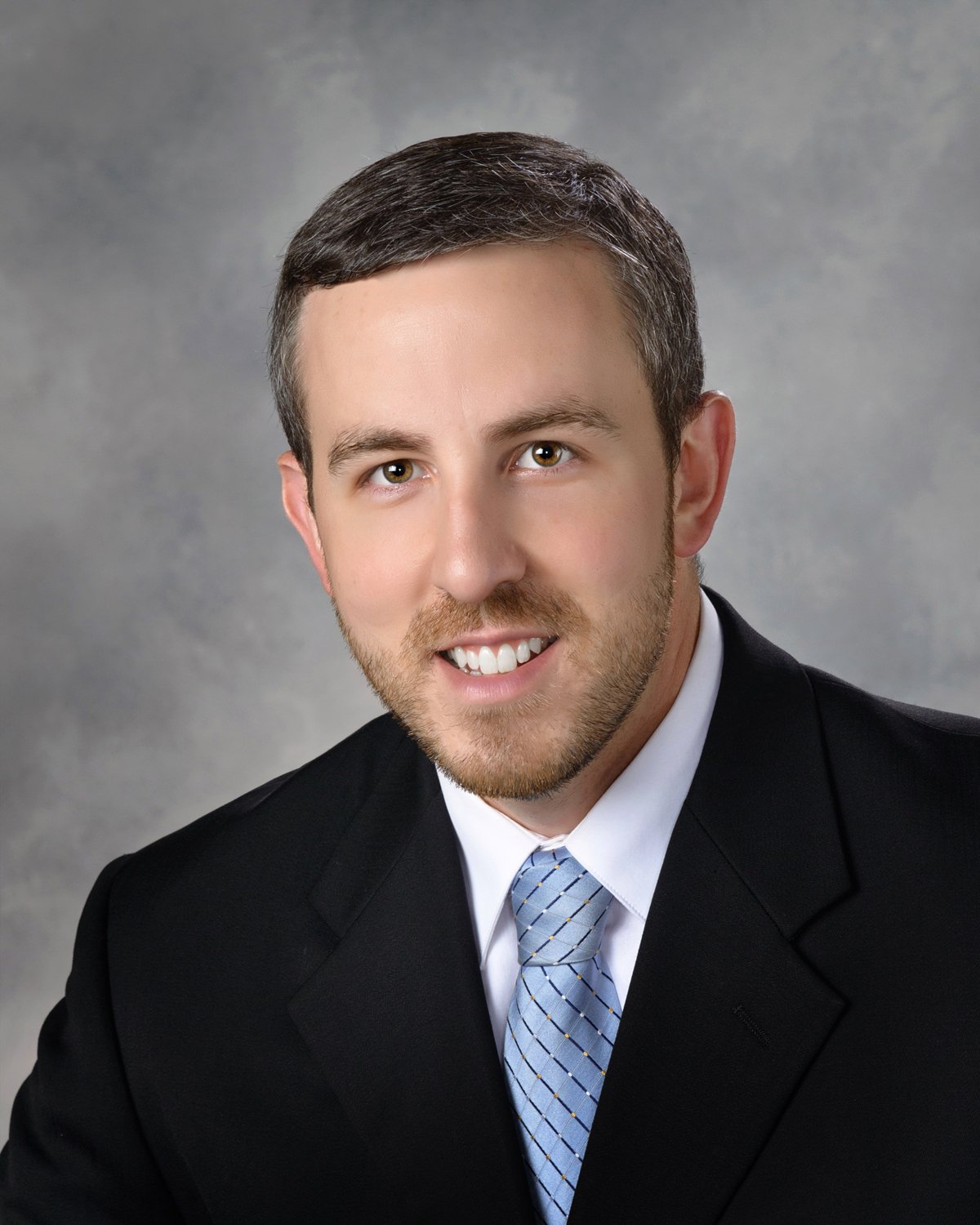 A professional headshot of a man with short brown hair and a beard, smiling, dressed in a black suit, white shirt, and a light blue tie, against a gray background.