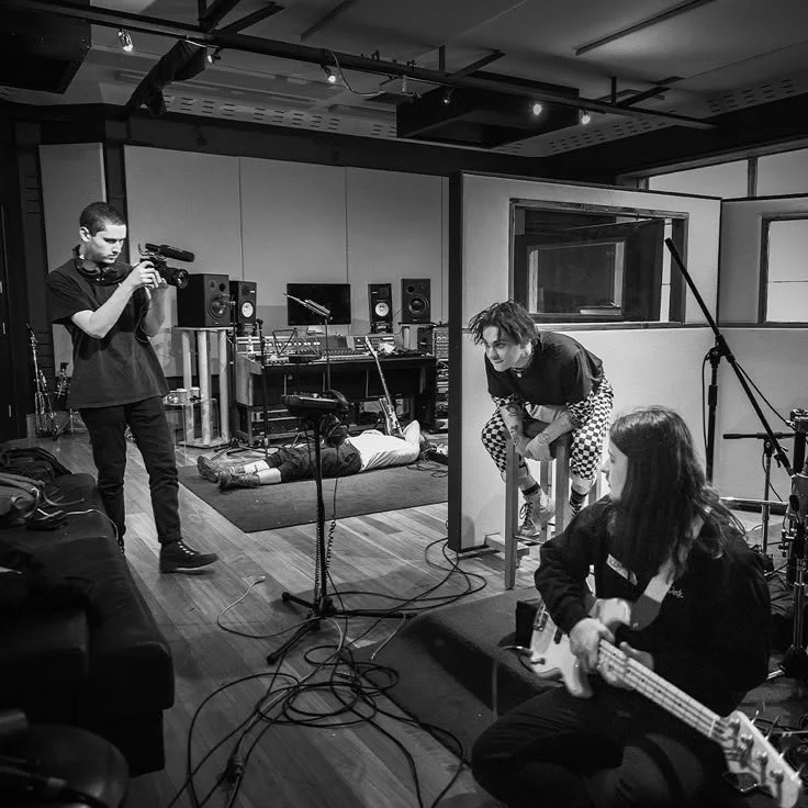 Músicos grabando juntos en un estudio, en blanco y negro: un cantante frente al micrófono y dos personas con instrumentos — acuerdos de colaboración y negociación entre artistas, Fuzz Legal.