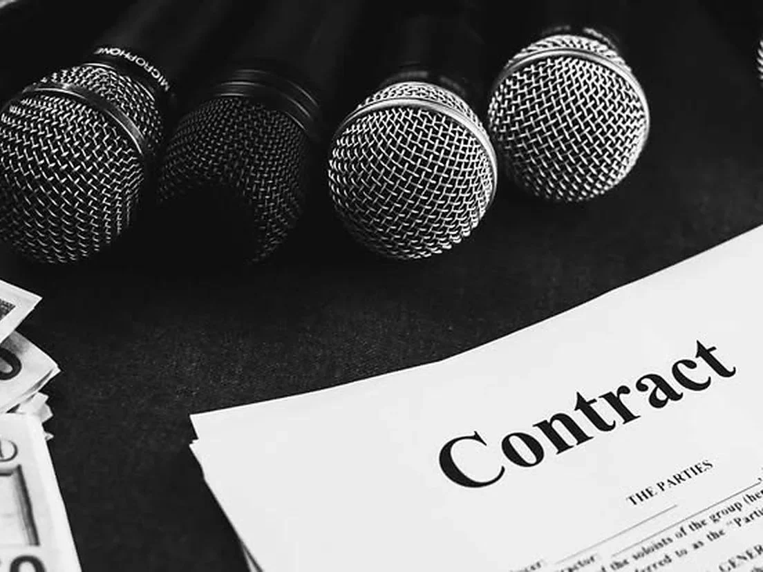 Cuatro micrófonos de mano en blanco y negro apoyados junto a un contrato impreso sobre una mesa — revisión de contratos discográficos, distribución y management, Fuzz Legal.