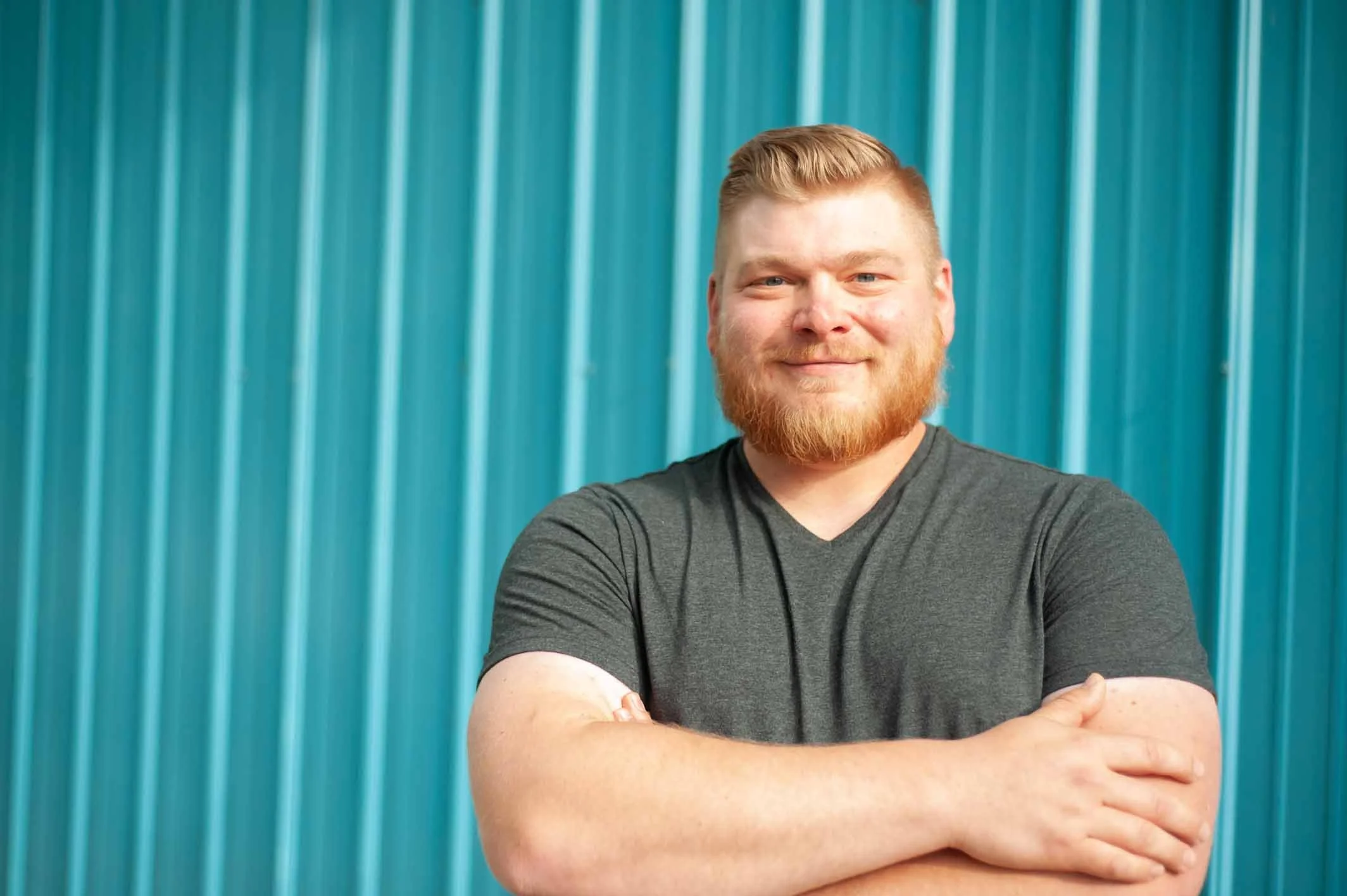 A young man with a beard and mustache, wearing a dark gray t-shirt, standing in front of a blue corrugated metal wall, smiling with his arms crossed.