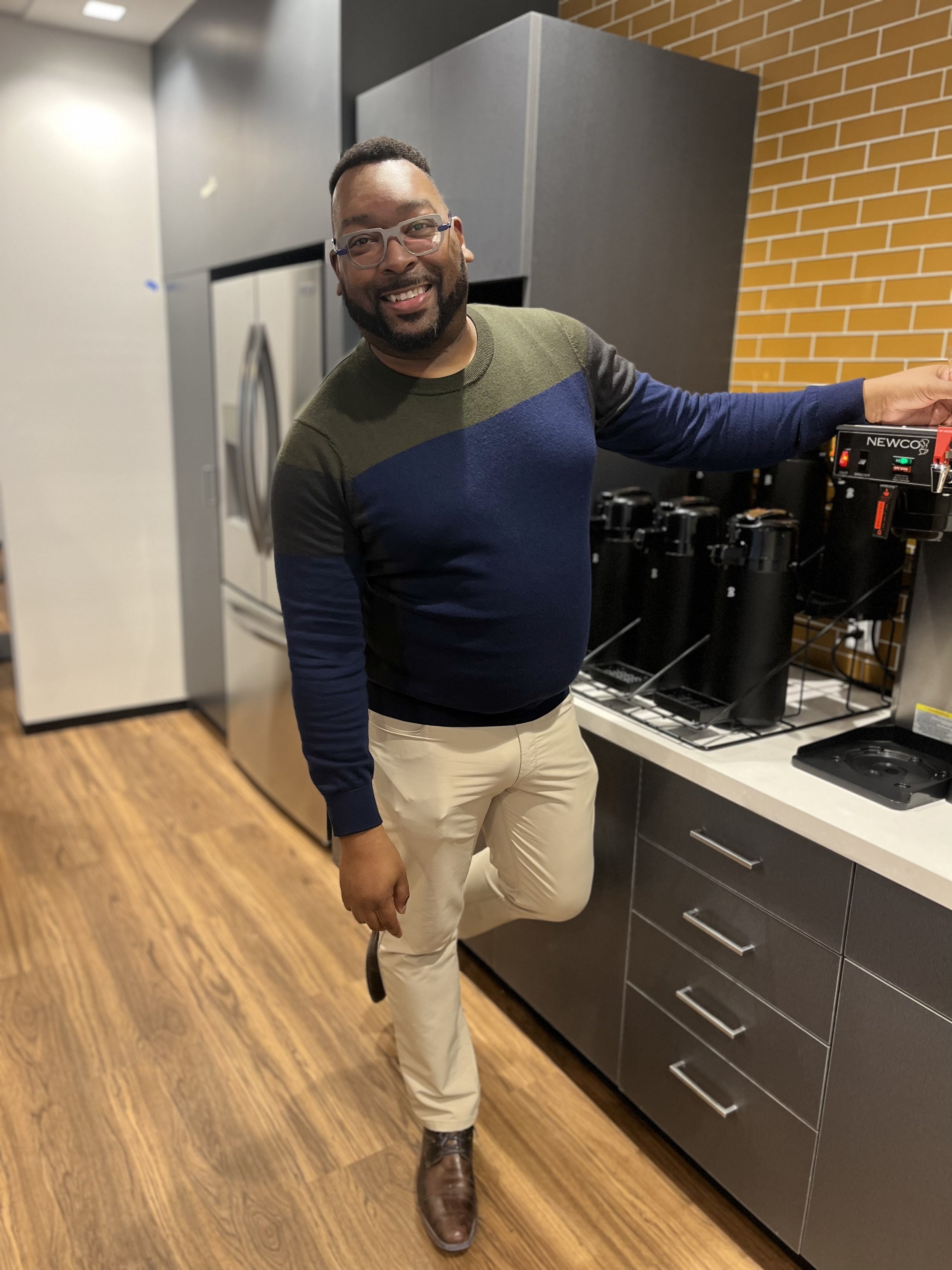 A man standing in a kitchen with one foot on the floor and the other resting on a kitchen counter. He is smiling, wearing glasses, a multi-colored sweater, beige pants, and brown boots. There are kitchen appliances including a refrigerator and a coffee maker in the background.