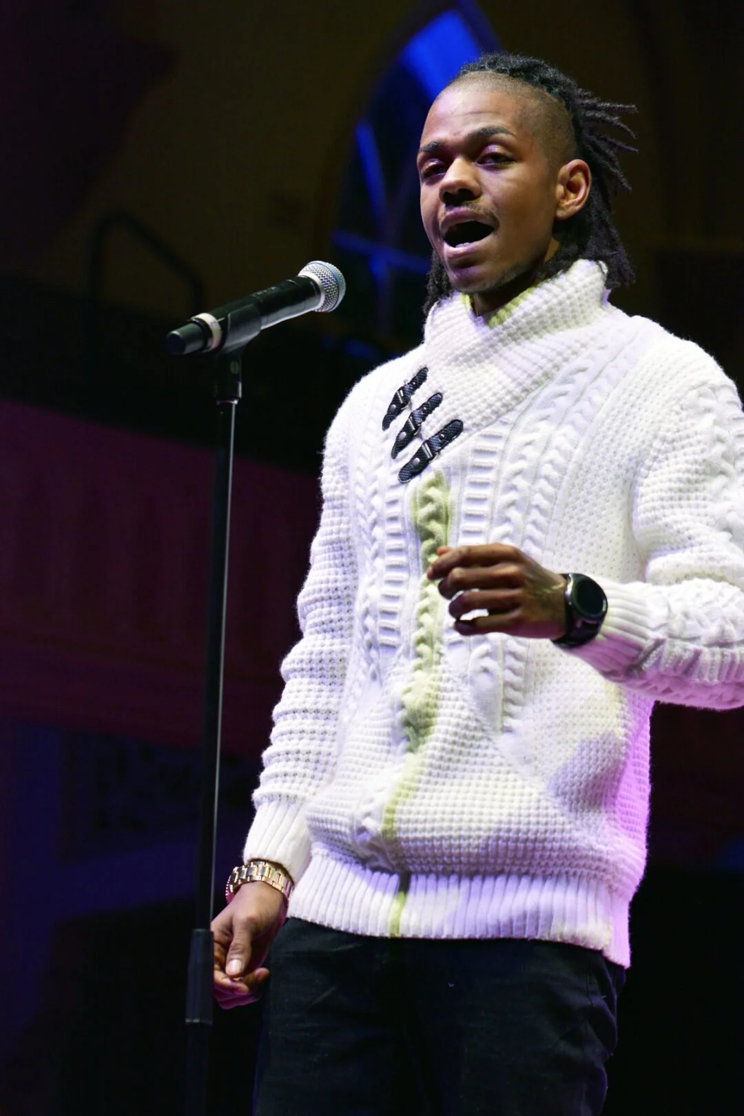 A man with tamed dreadlocks speaking or singing into a microphone on stage, wearing a cream-colored knitted jacket and a watch.