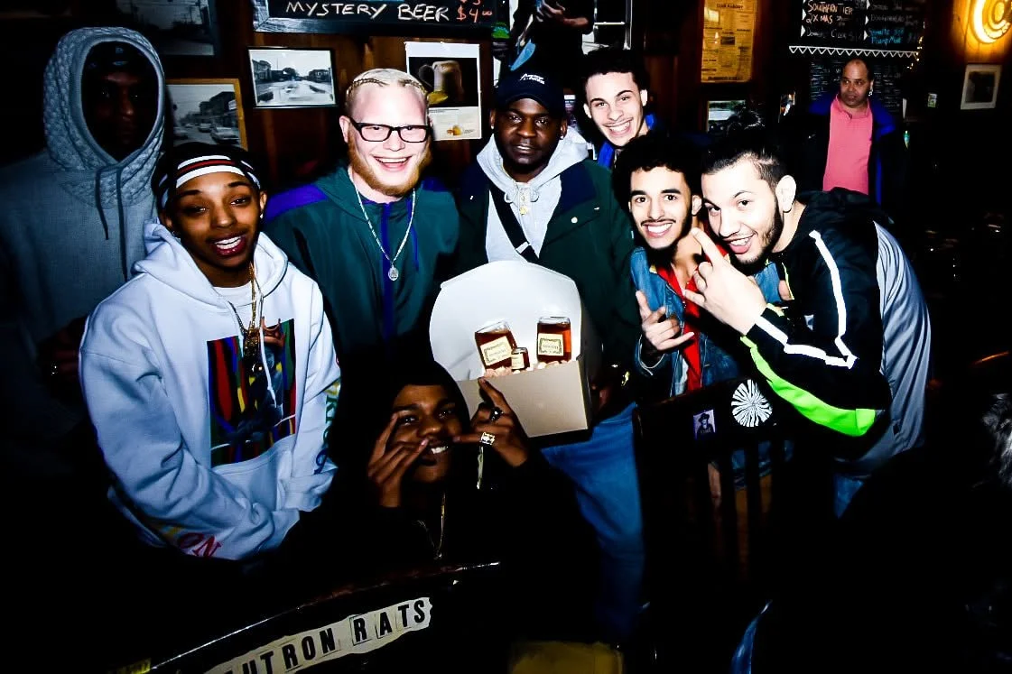 A group of eight diverse young people smiling and posing together inside a restaurant or bar. They are gathered around a table, with some holding a box that contains bottles of Jack Daniel's whiskey. The background shows framed pictures and a chalkboard menu.