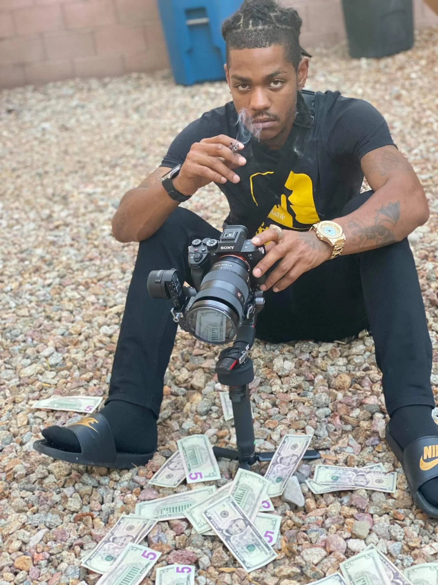 A young man with tattoos, dressed in black, sitting on a gravel surface with scattered cash, holding a camera, and smoking a cigarette.