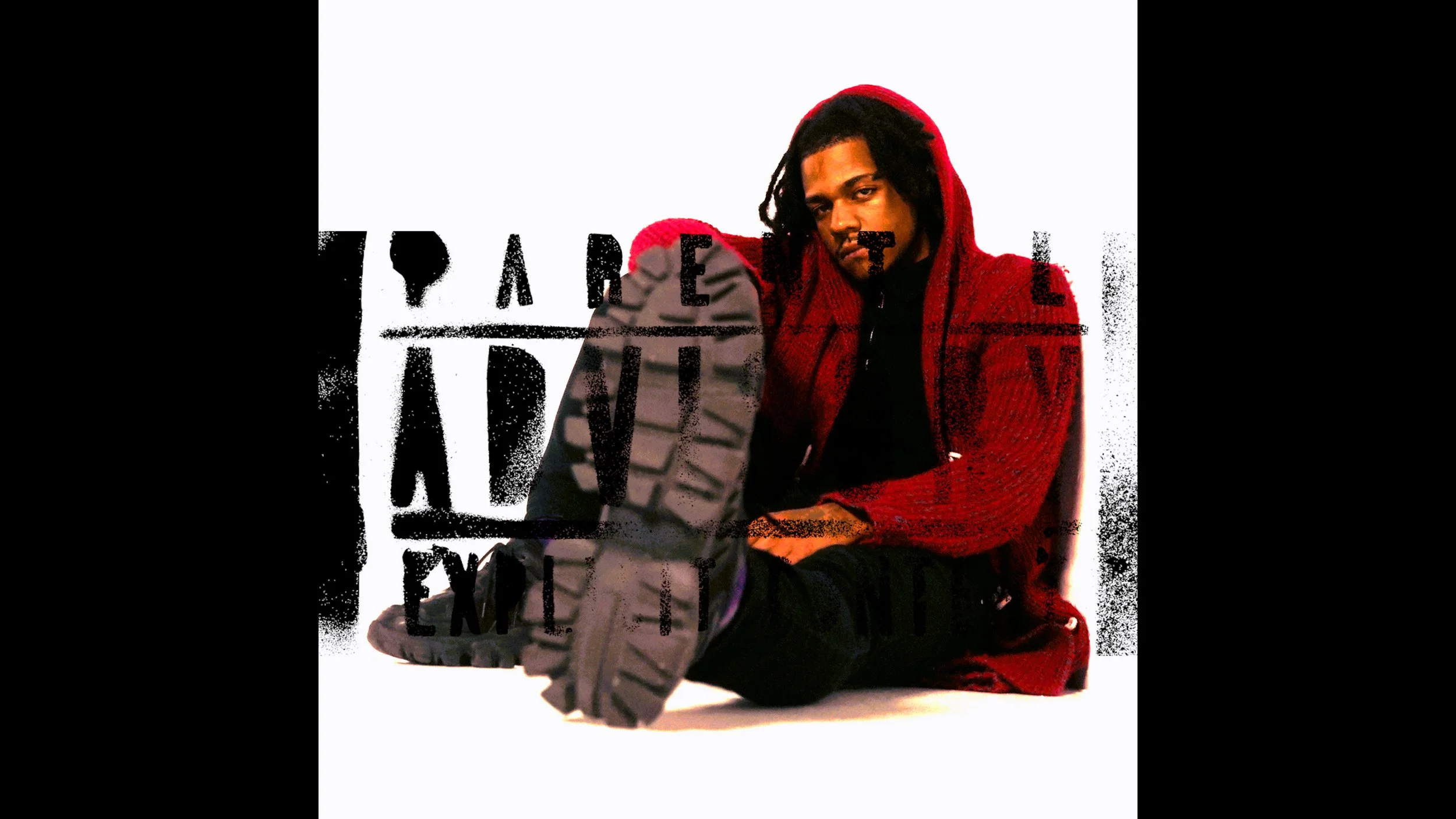 A young man with dreadlocks wearing a red hoodie and black clothing, sitting against a white background with black graffiti text partially visible.