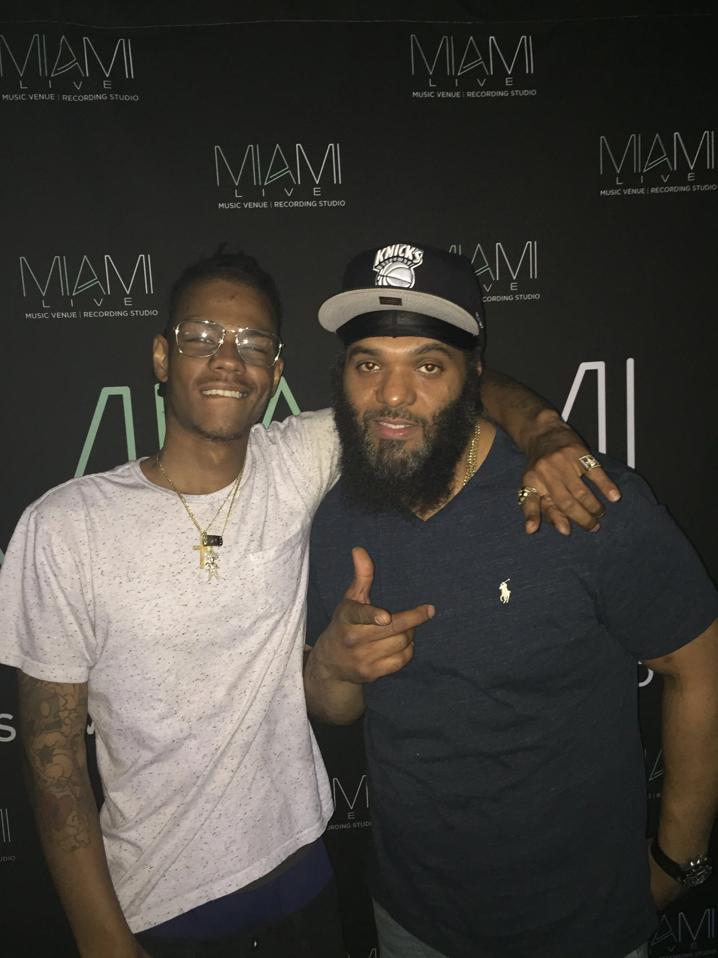 Two men posing for a photo in front of a black background with the text 'Miami Live' and 'Music Venue | Recording Studio' repeated across it. One man is wearing glasses and a white T-shirt, the other is wearing a black cap with a New York Knicks logo and a dark shirt, and they are standing close together with their arms around each other.