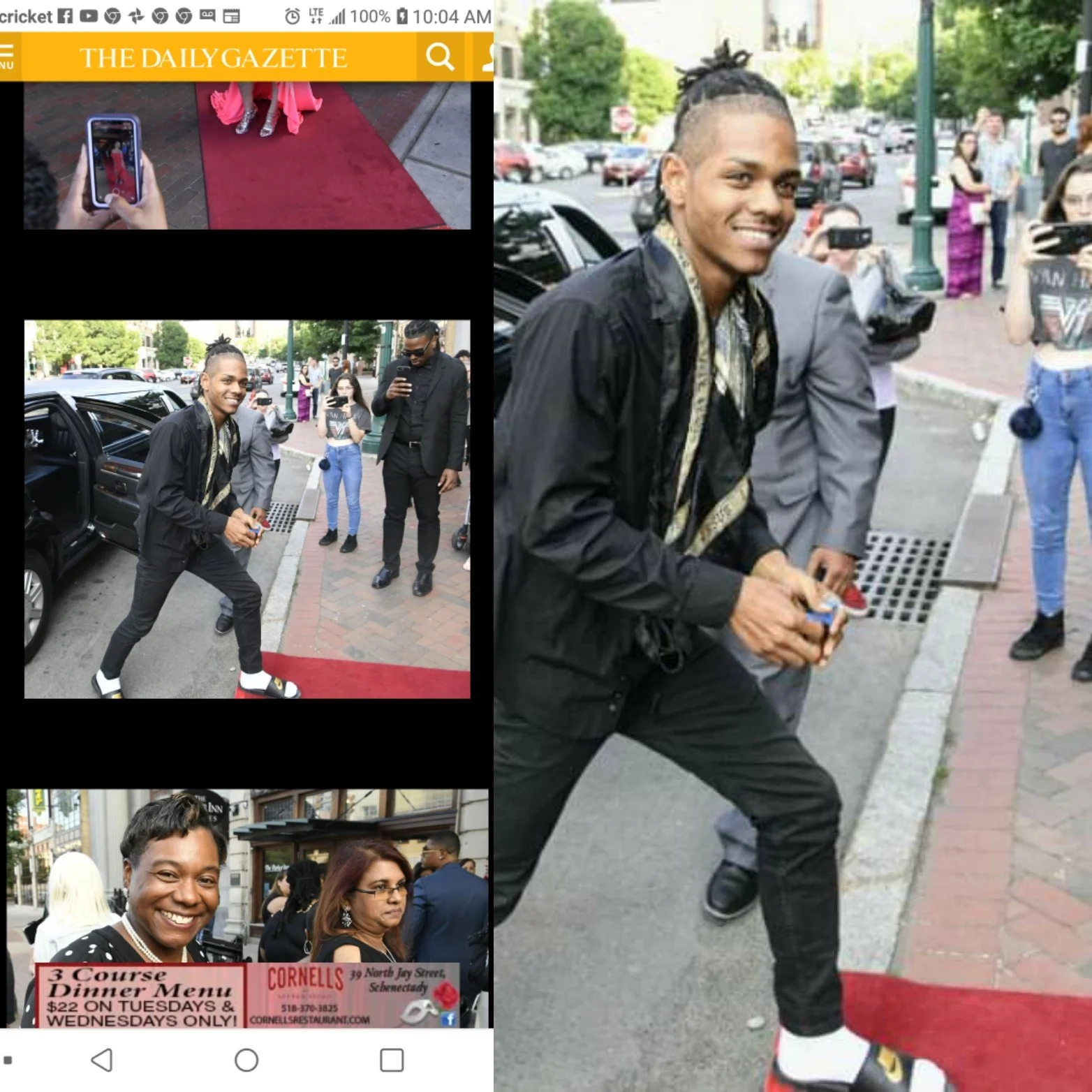 A man with dreadlocks tied up, wearing a black jacket, black pants, and sneakers, smiling and standing on a city sidewalk while holding something in his hands, with several people and parked cars in the background.