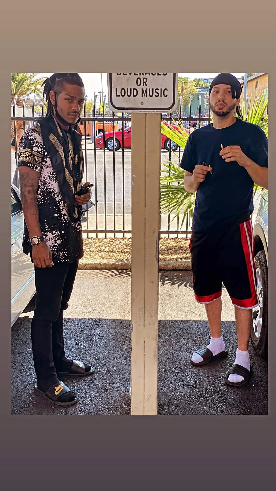 Two young men standing outdoors near a fence, with a sign above them that reads 'BEVERAGES OR LOUD MUSIC', and parked cars in the background.