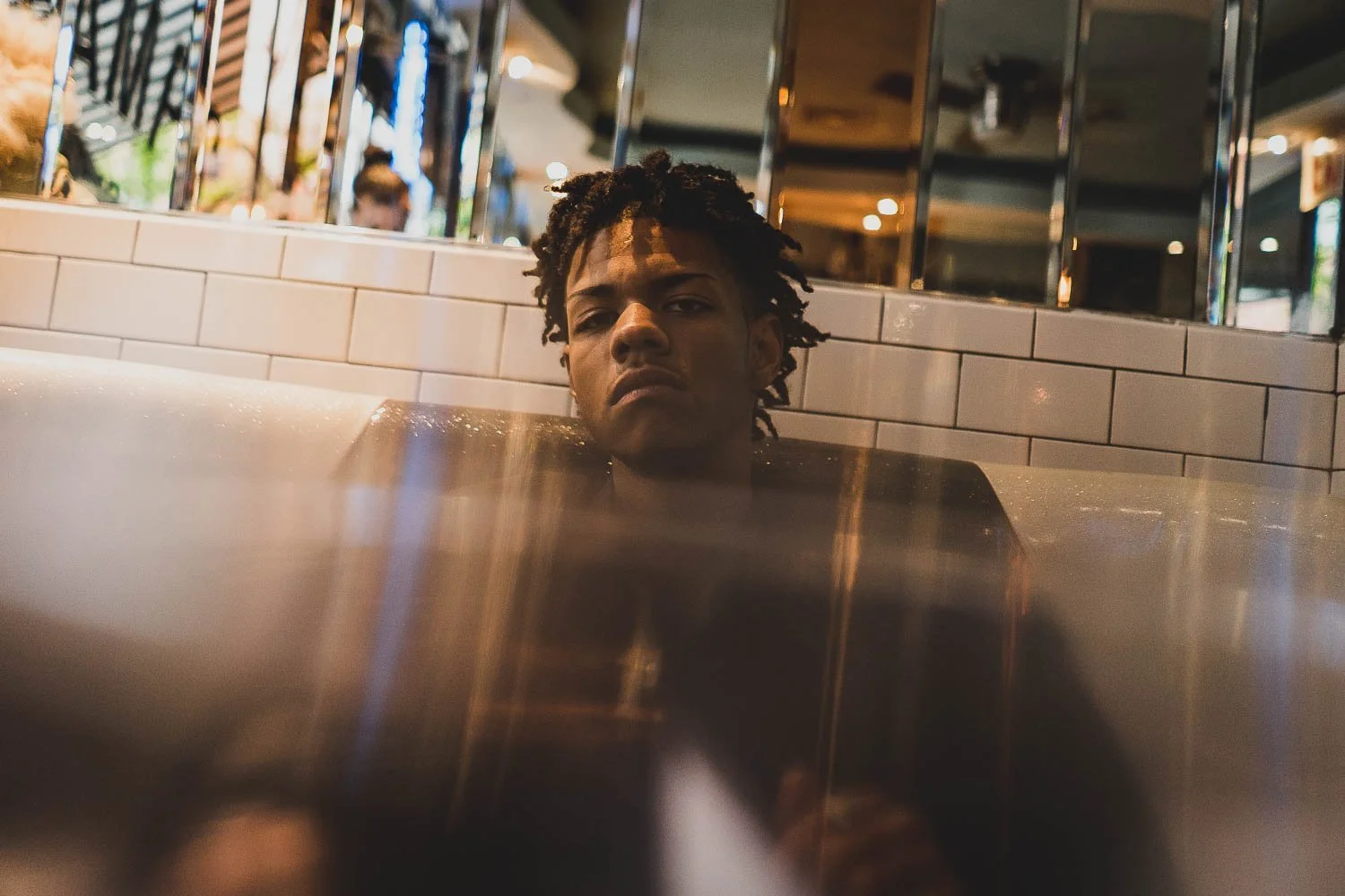 A young man with dreadlocks sitting in a bathtub with a serious expression in a modern, dimly lit restaurant or cafe.