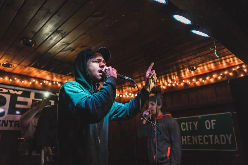 A man singing into a microphone on stage with string lights overhead and a city of Henectady sign in the background.
