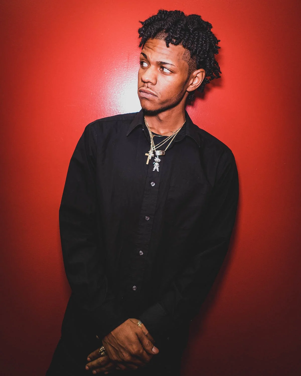 Young man with curly hair wearing a black shirt and layered necklaces, standing against a red wall with a thoughtful expression.
