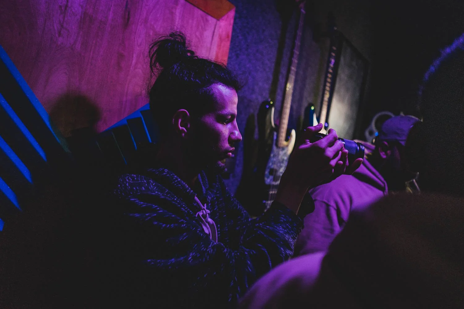 Person with dark hair in a bun, wearing a patterned hoodie, sitting against a wooden wall with guitars hanging behind, holding a camera, in a dimly lit room with purple and blue lighting.