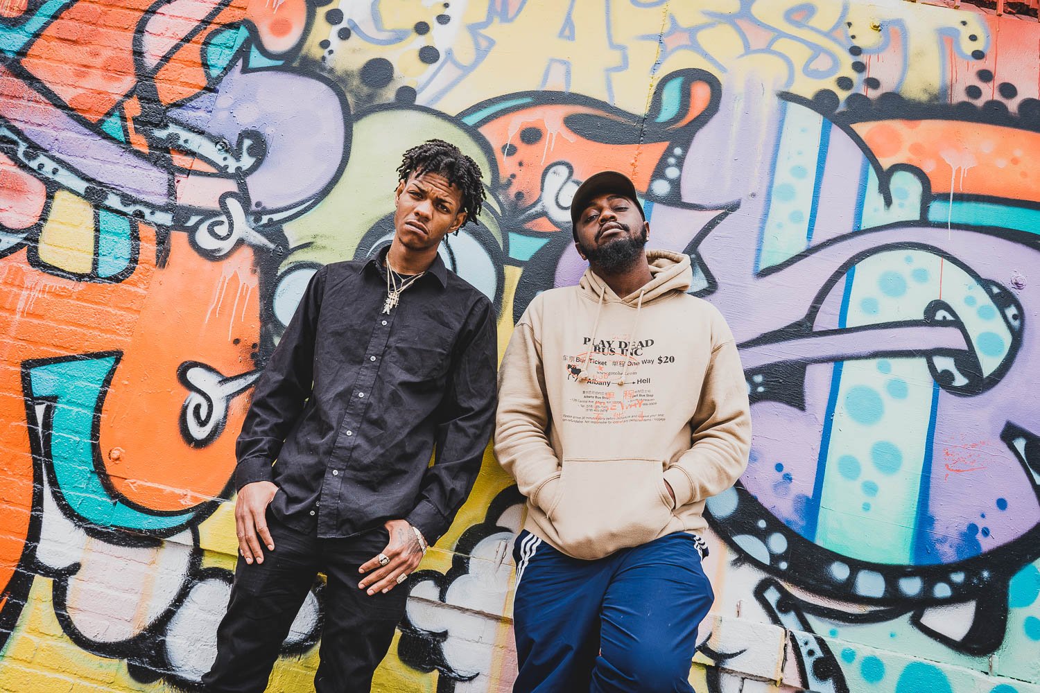 Two young men standing in front of a colorful graffiti wall, looking at the camera with serious expressions.