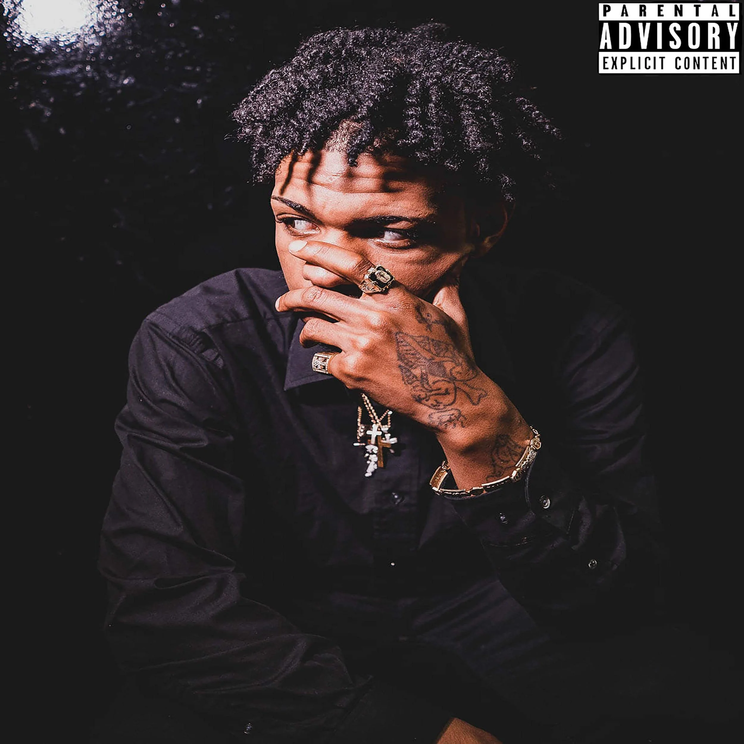 A man with curly black hair covering part of his face, wearing rings and a chain necklace, appears to be covering his face with his hand, and is dressed in a dark jacket, posing against a dark background.