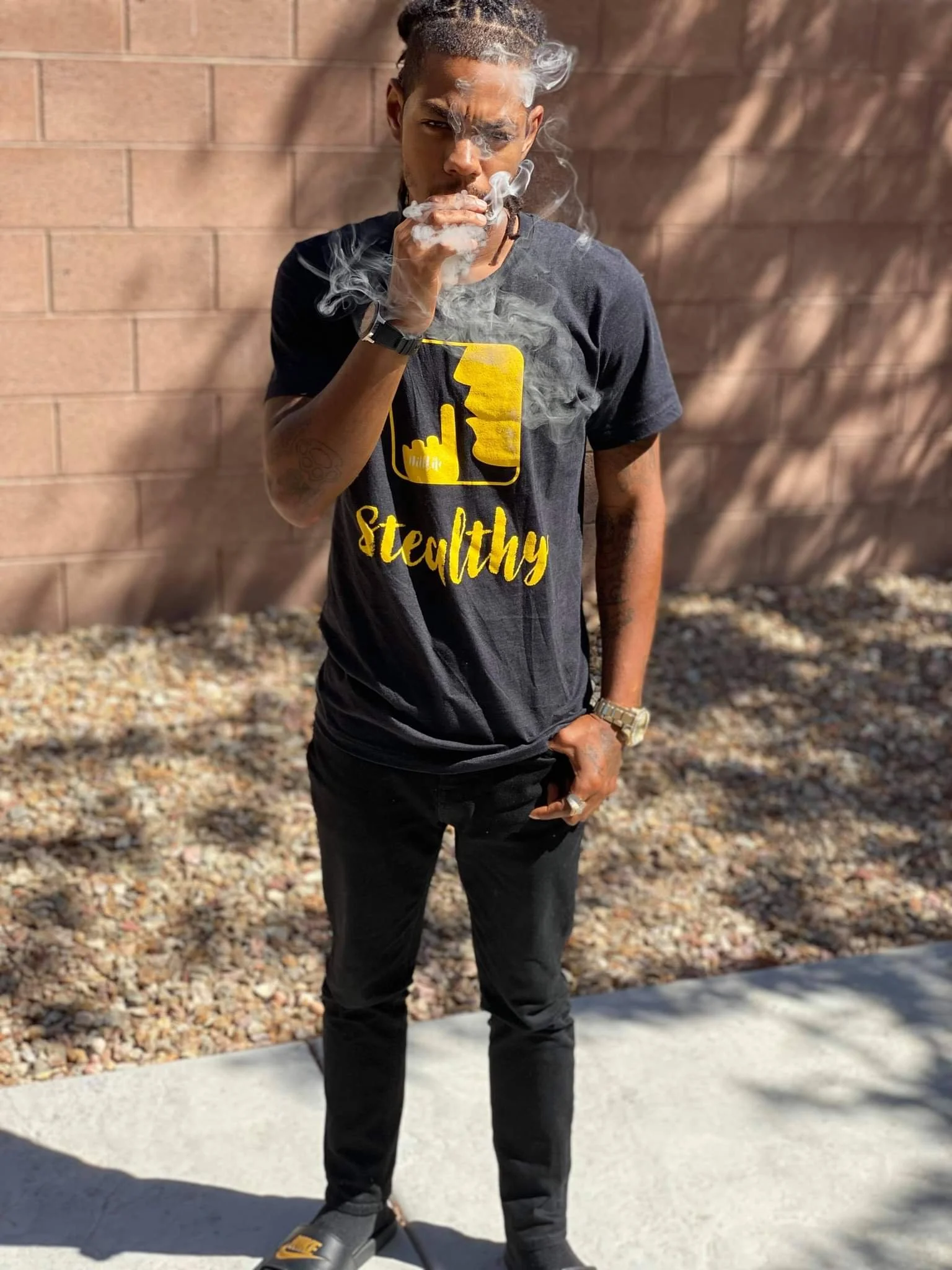 A young man with dreadlocks and tattoos, wearing a black t-shirt with yellow graphics and text that reads 'Stelthy,' standing outdoors against a brick wall, smoking with smoke around his face.