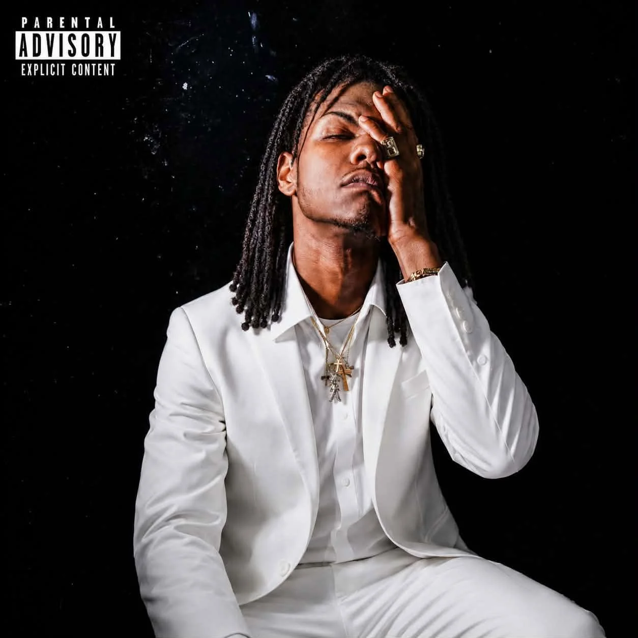 A man with long dreadlocks, wearing a white suit and multiple jewelry pieces, faces a black background with one hand covering part of his face.