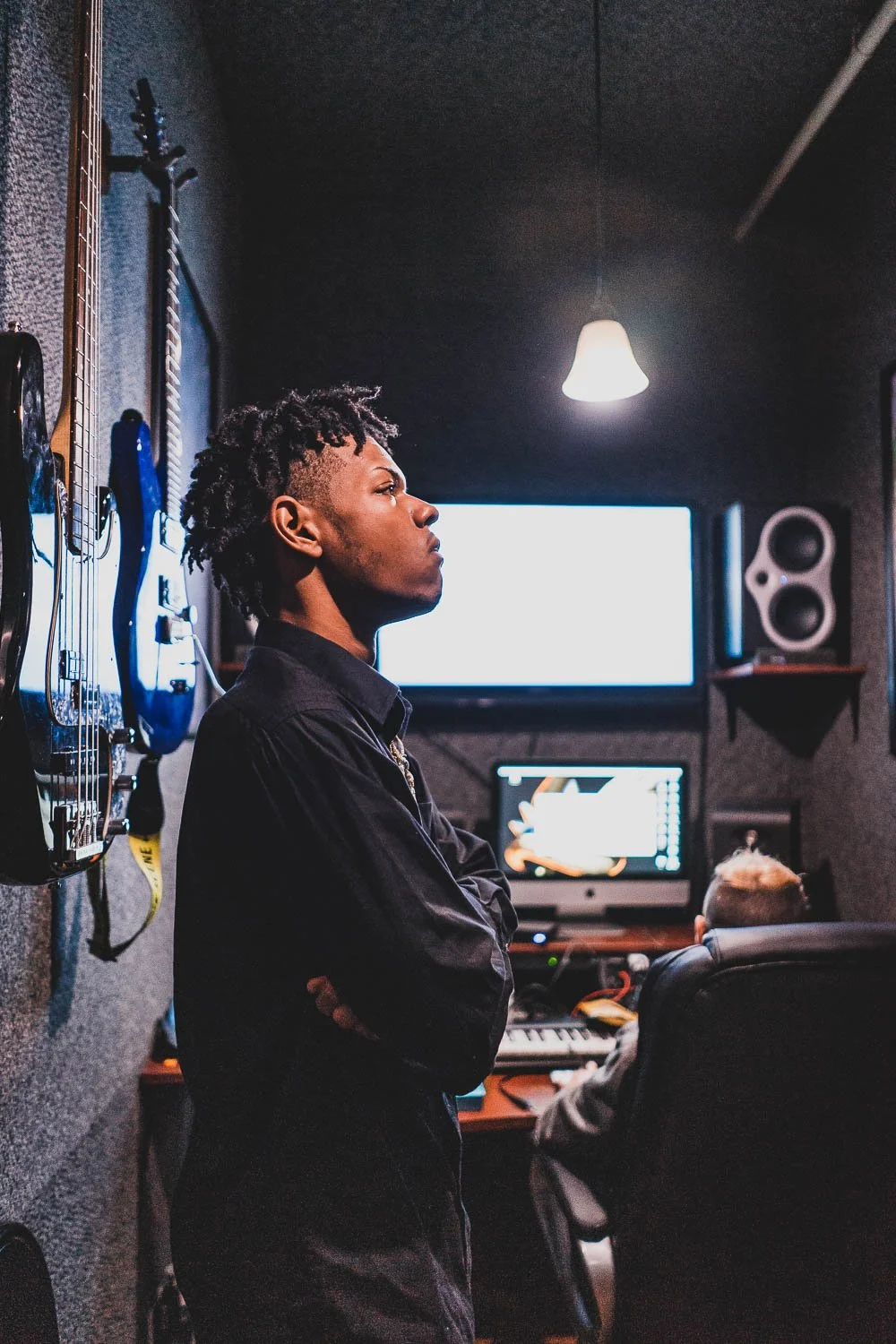 A young man with dreadlocks stands in a music studio, facing a wall with several guitars hanging, while a person with short blonde hair works at a computer in the background.