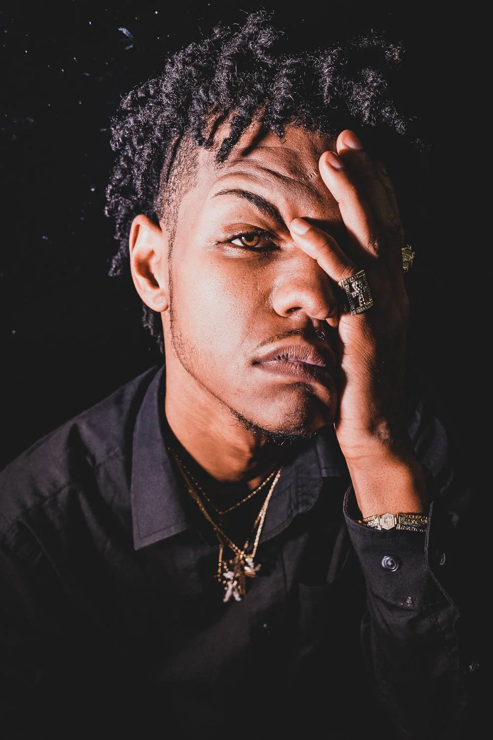 A young man with curly hair and gold jewelry covers one eye with his hand, set against a dark background.