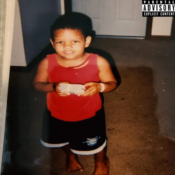A young boy with short curly hair, wearing a red sleeveless shirt and black shorts, standing in a room with a carpeted floor, holding a small white object in his hands, and looking at the camera.
