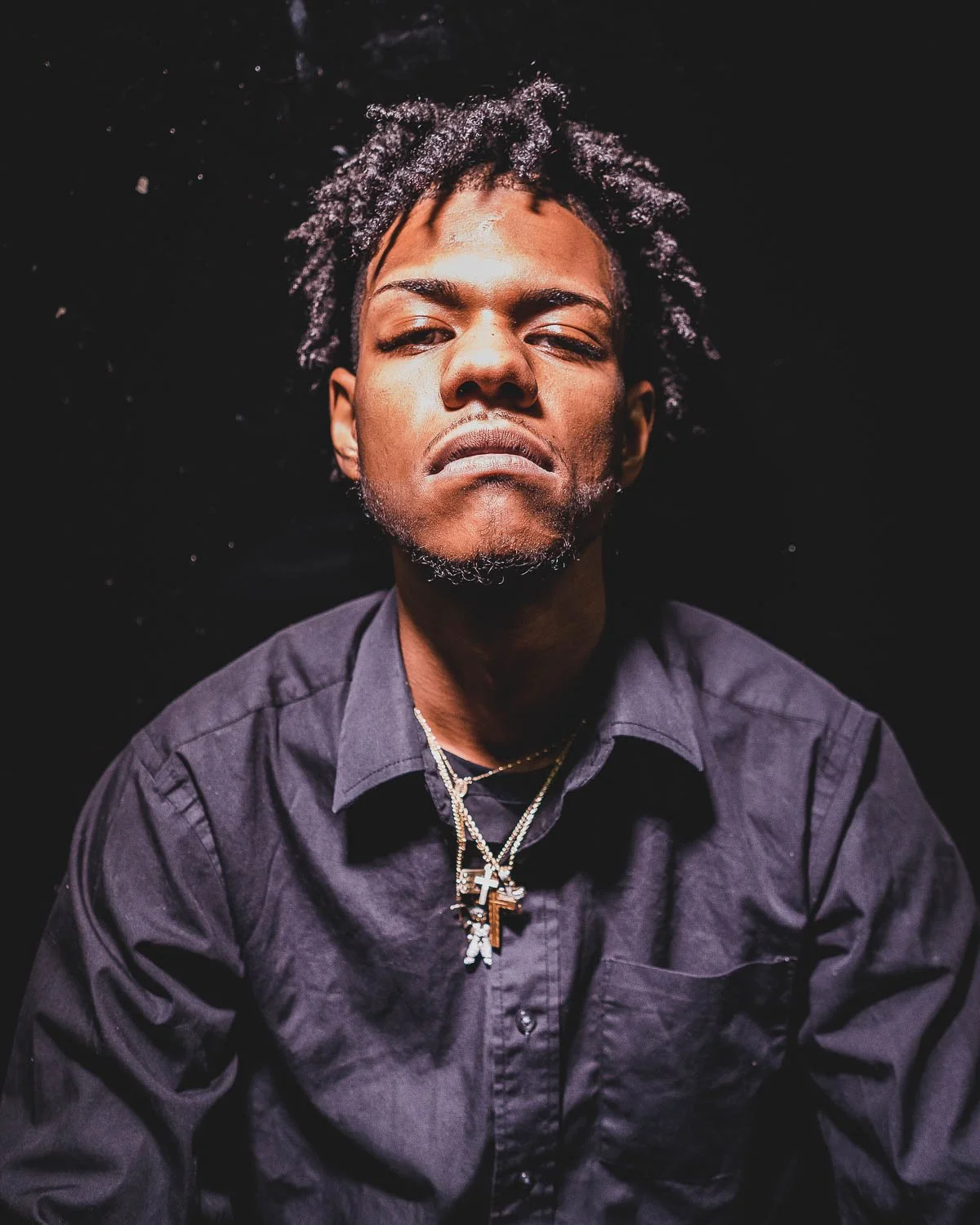 A man with dark skin and curly hair with white tips, wearing a dark shirt and layered necklaces, looking directly at the camera with a serious expression in a dark setting.