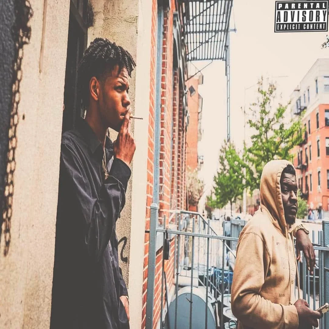 Two young men standing outside on a city street, one smoking and the other holding a phone, with buildings and trees in the background.