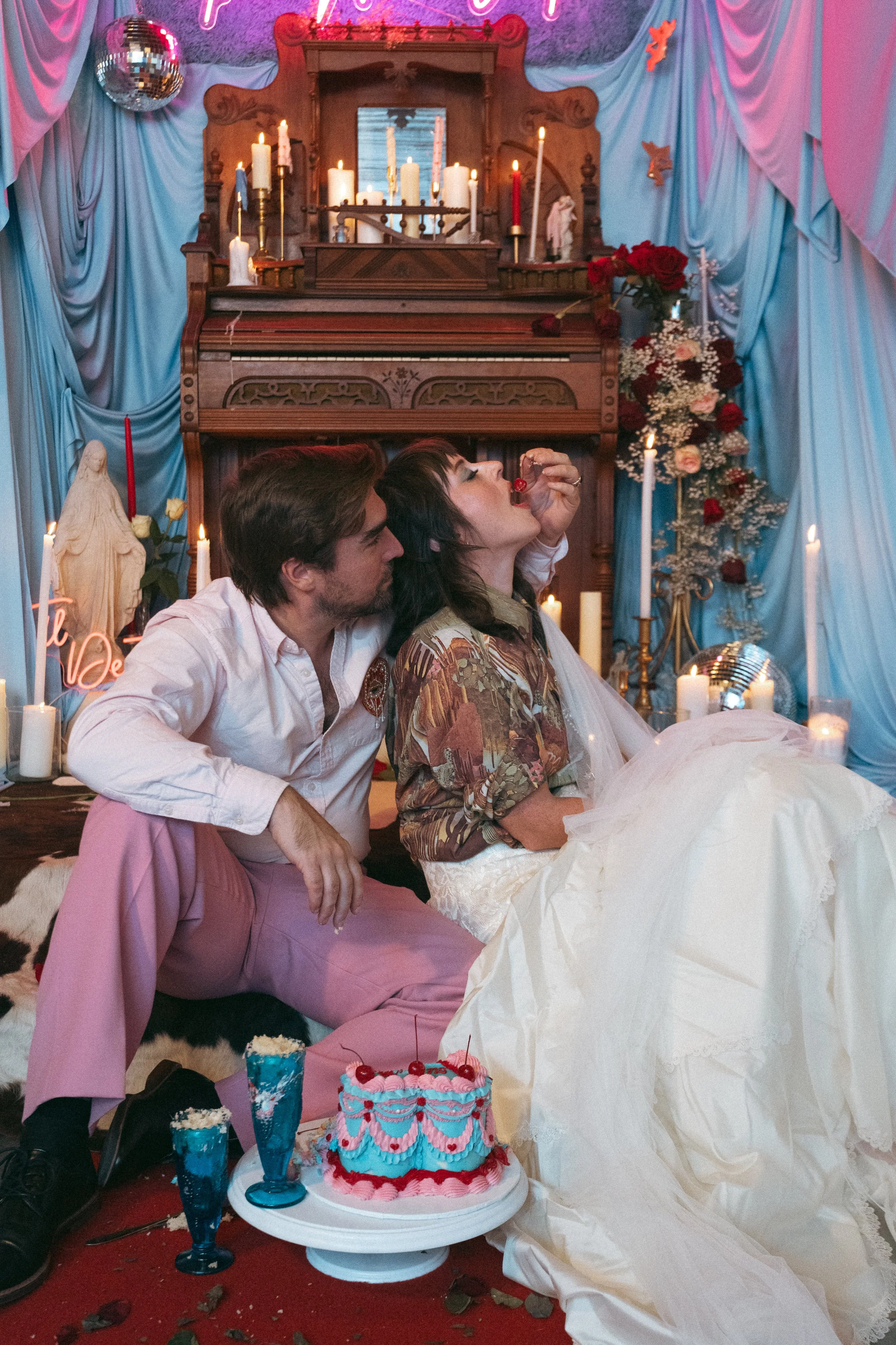 A couple celebrating a birthday with a cake, party decorations, candles, and a room decorated with blue curtains and flowers.