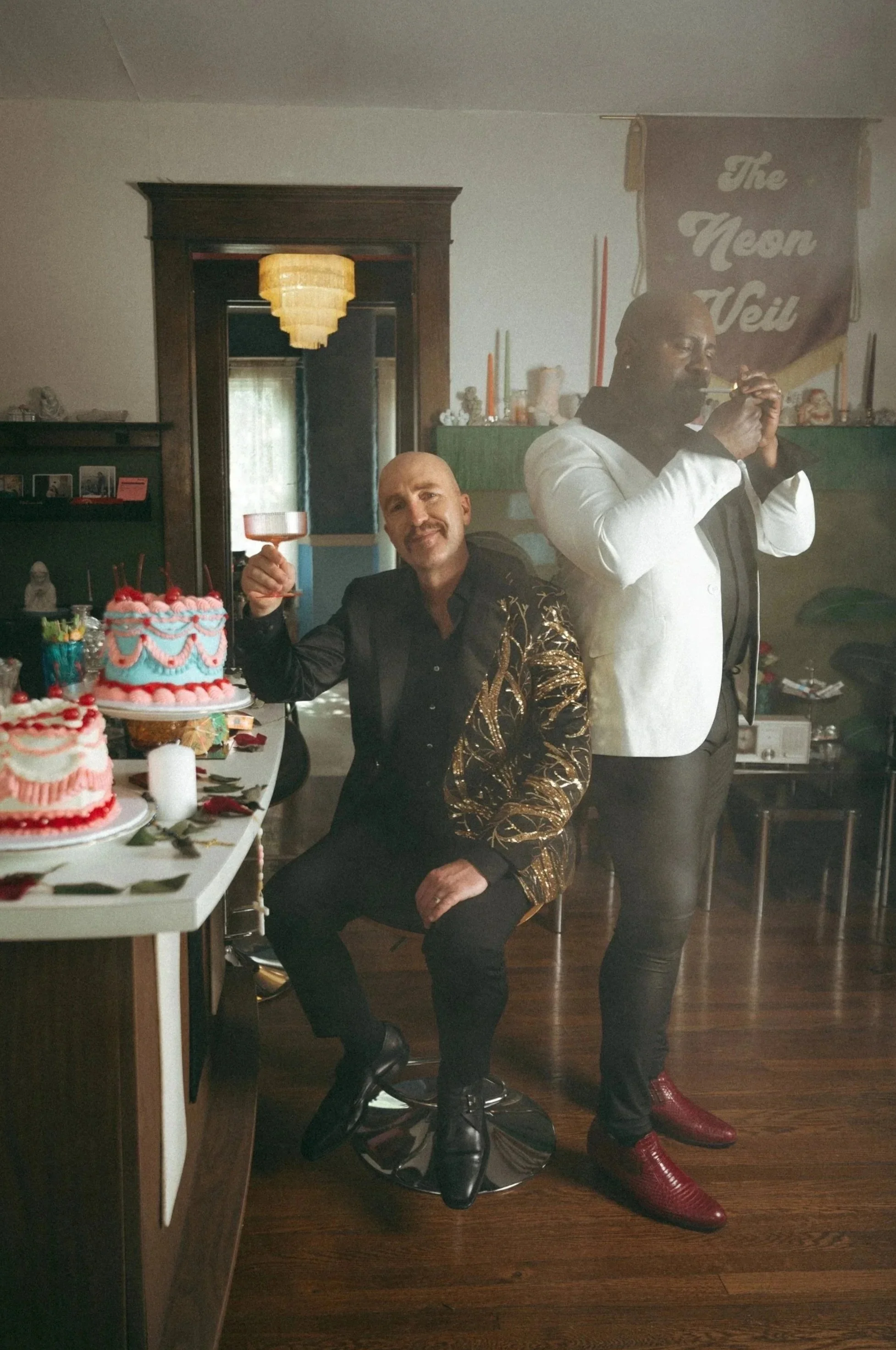 Two men celebrating at a birthday party with cakes, decorations, and a banner that reads 'The Neen Veil'. One man with a bald head and mustache is sitting and holding a glass, while the other man with glasses and darker skin is standing, about to blow out birthday candles on a cake.