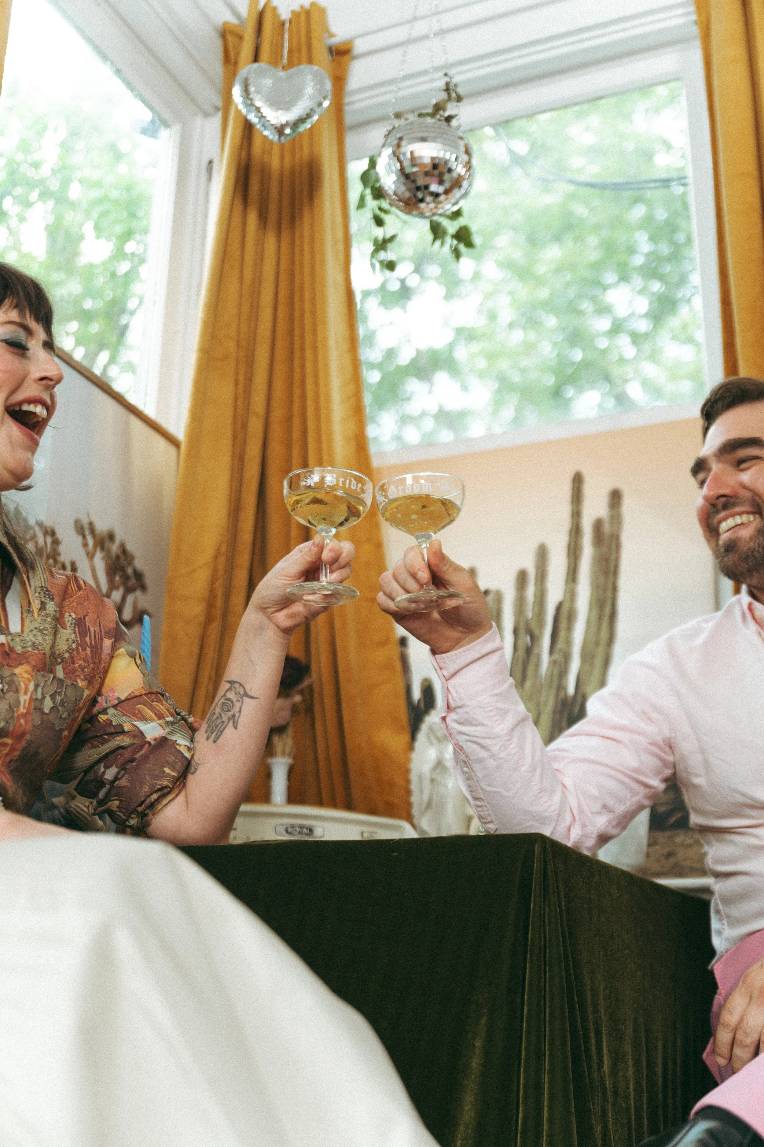 Two women and one man are celebrating with champagne glasses, raising their glasses with a toast in a cozy indoor space. They are smiling and appear cheerful, with the background showing yellow curtains, large windows with greenery outside, and disco ball decorations hanging from the ceiling.