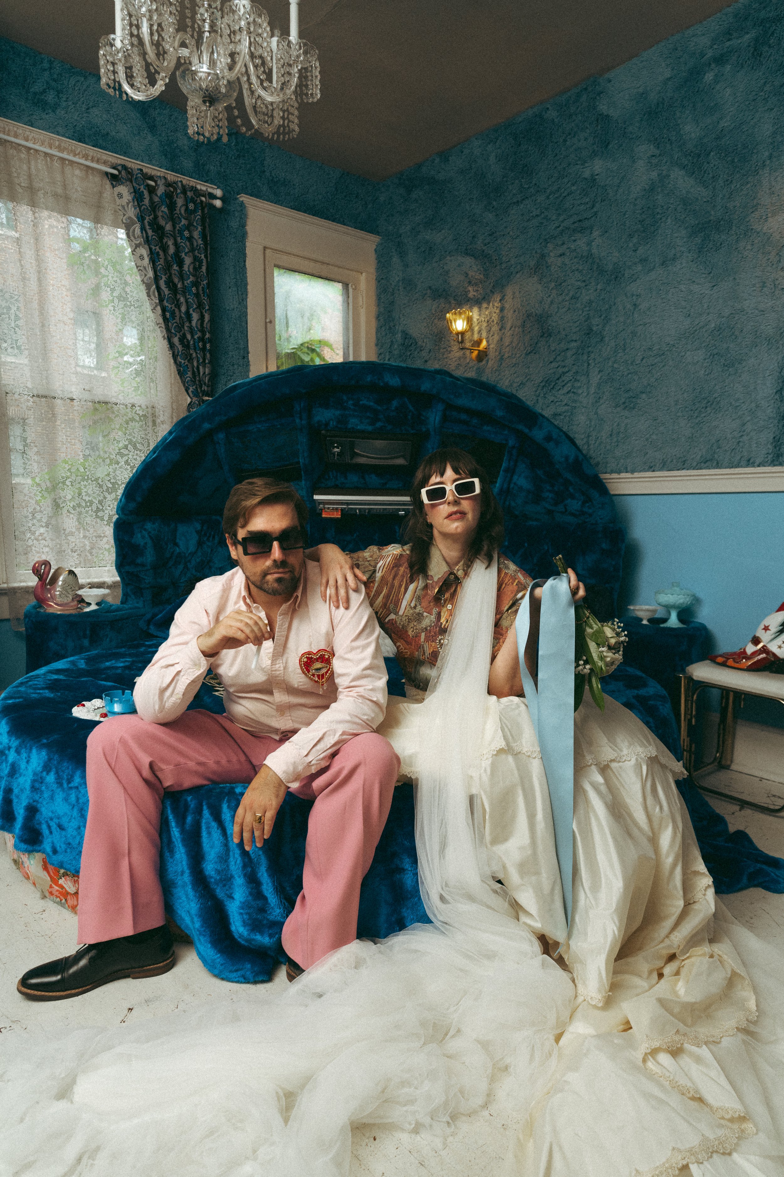 Two people sitting on a bed in a room decorated with blue walls, a chandelier, and lace curtains. The man wears sunglasses, a pink shirt, and pink pants, while the woman wears sunglasses, a patterned shirt, and a white dress with a blue ribbon. There are various items on the bed, including a gift bag, flowers, and a small figurine.
